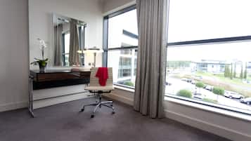 A hotel room with a large window, a desk with a chair, a mirror, and a vase with a plant.