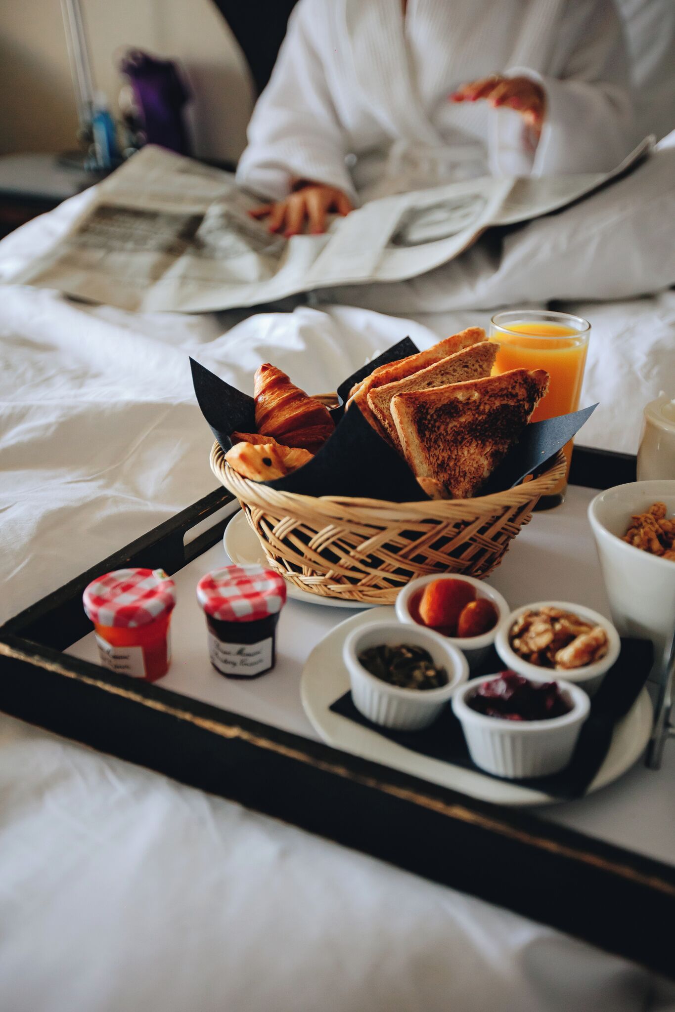 Bandeja de desayuno con tostadas, croissants y un vaso de jugo de naranja sobre una cama.