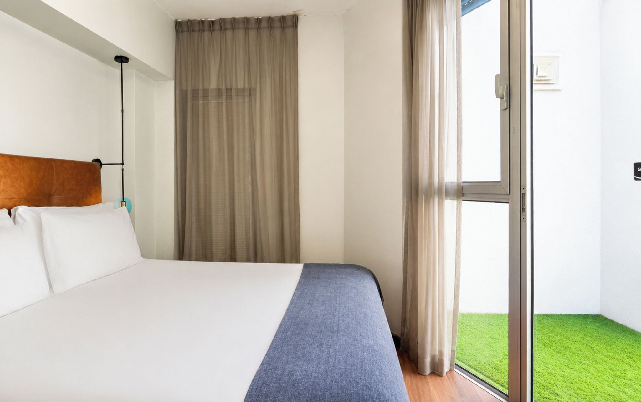 Habitación de hotel con cama, cabecero, ventana con cortinas y vista a un césped verde.