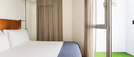 Habitación de hotel con cama, cabecero, ventana con cortinas y vista a un césped verde.