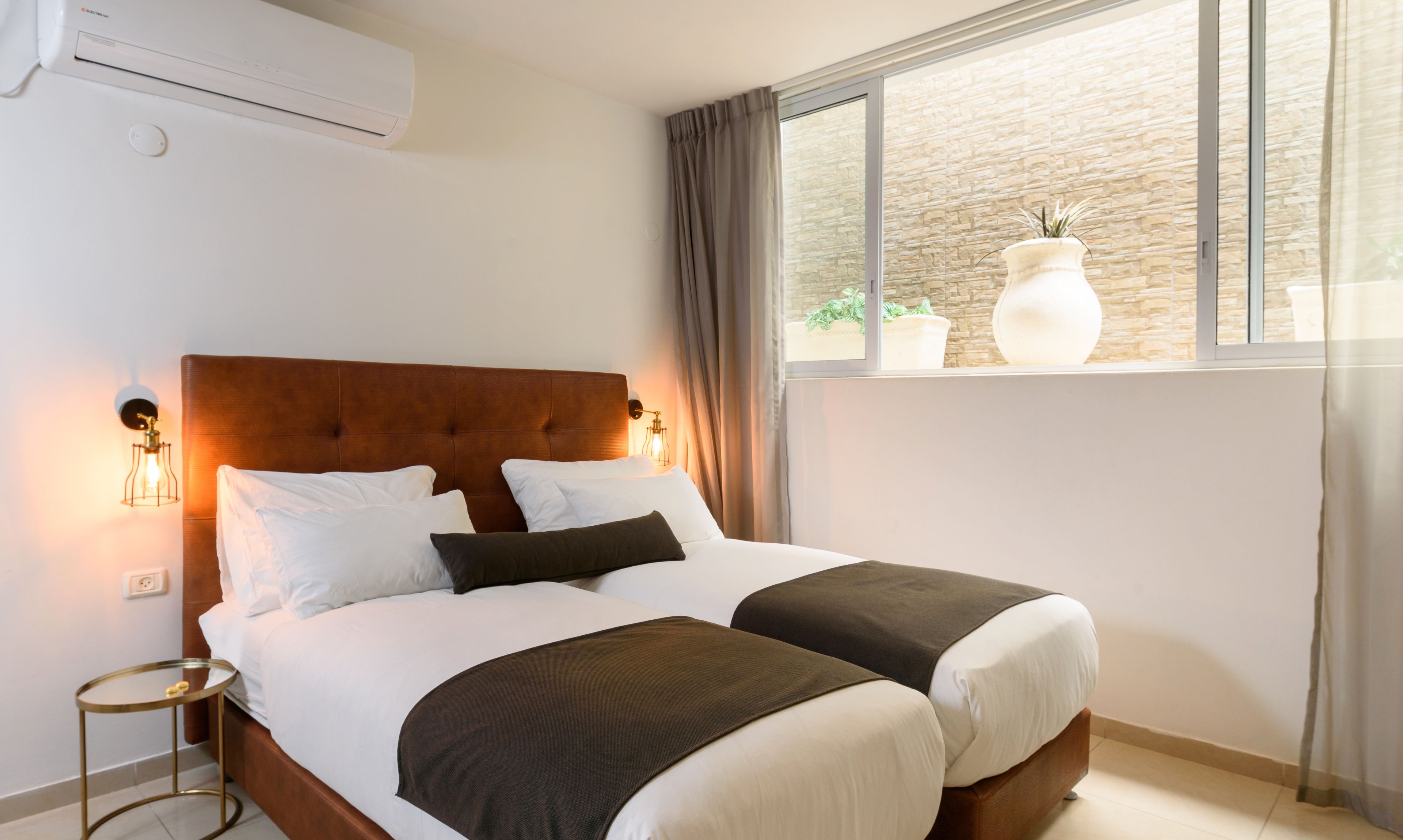 A double bed with a wooden headboard, two bedside tables, a wall-mounted air conditioner, and a window with curtains.