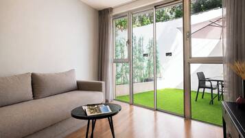 A modern living room with a sofa, a small round table with a magazine, and a view of a green lawn through large glass doors.