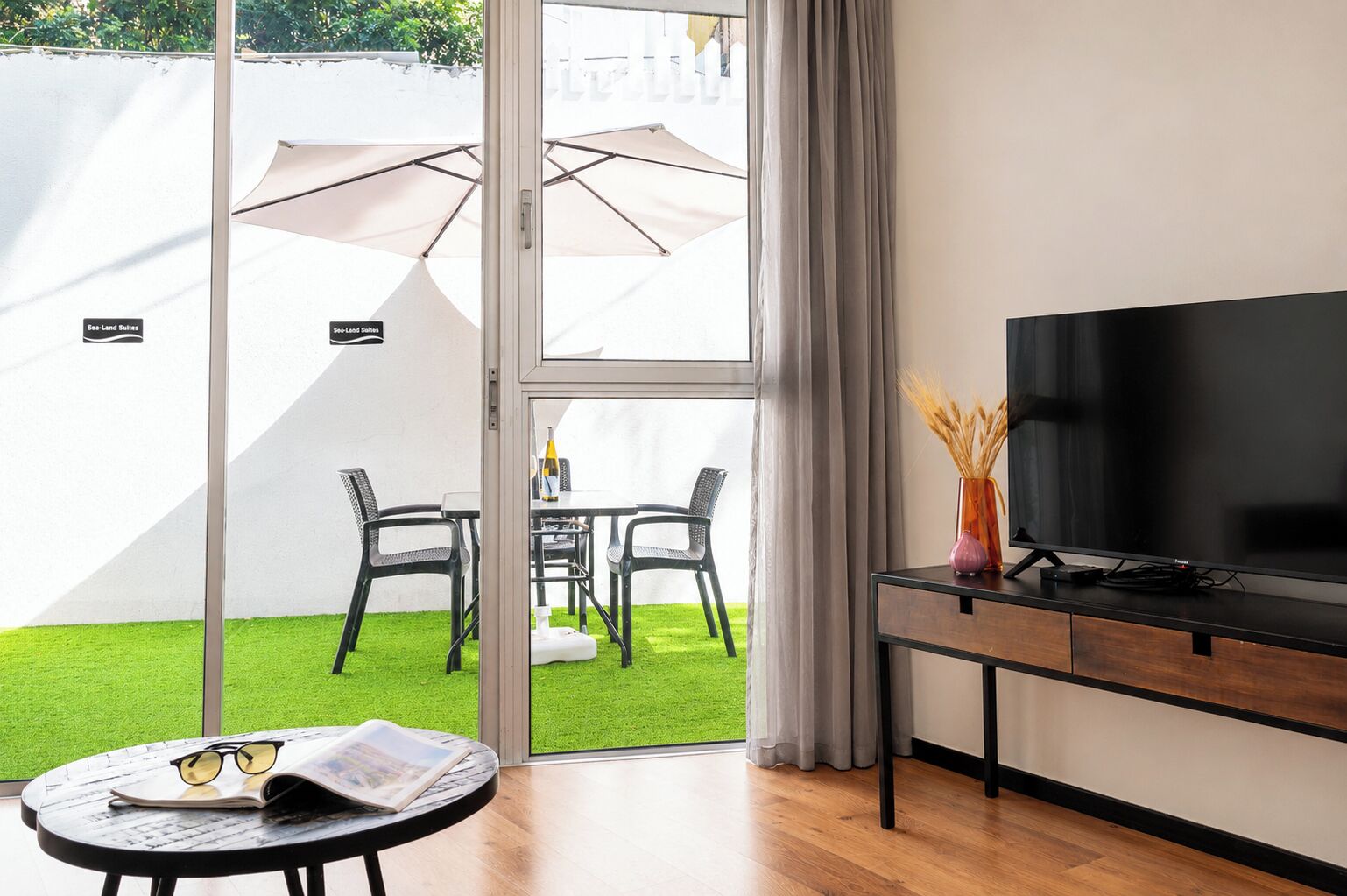 A room with a TV, a table with glasses and a magazine, a chair, and a view of a garden with a table and chairs outside.