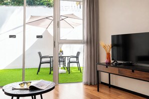 A room with a TV, a table with glasses and a magazine, a chair, and a view of a garden with a table and chairs outside. - Sea Land Suites - by Echo Hotels (Tel Aviv)