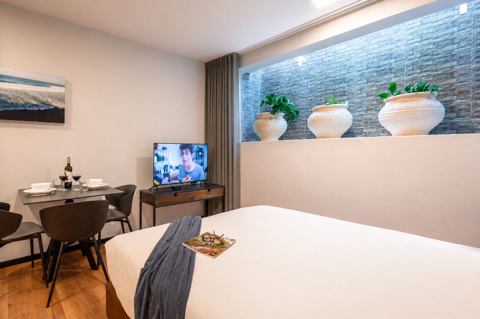 Quarto de hotel moderno com cama, escrivaninha com televisão e janela com vasos decorativos.