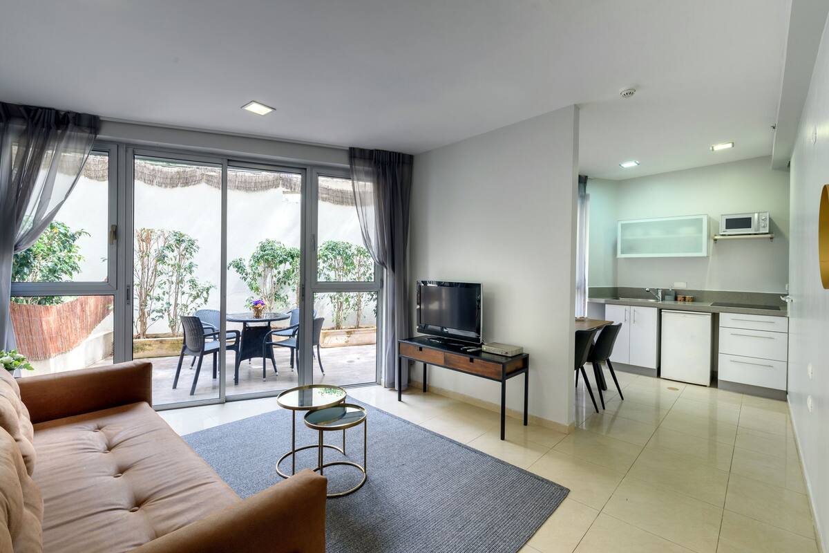 A modern living room with a sofa, a coffee table, and a TV. There is a sliding glass door leading to an outdoor patio with a table and chairs. A kitchen area is visible with a microwave and cabinets.