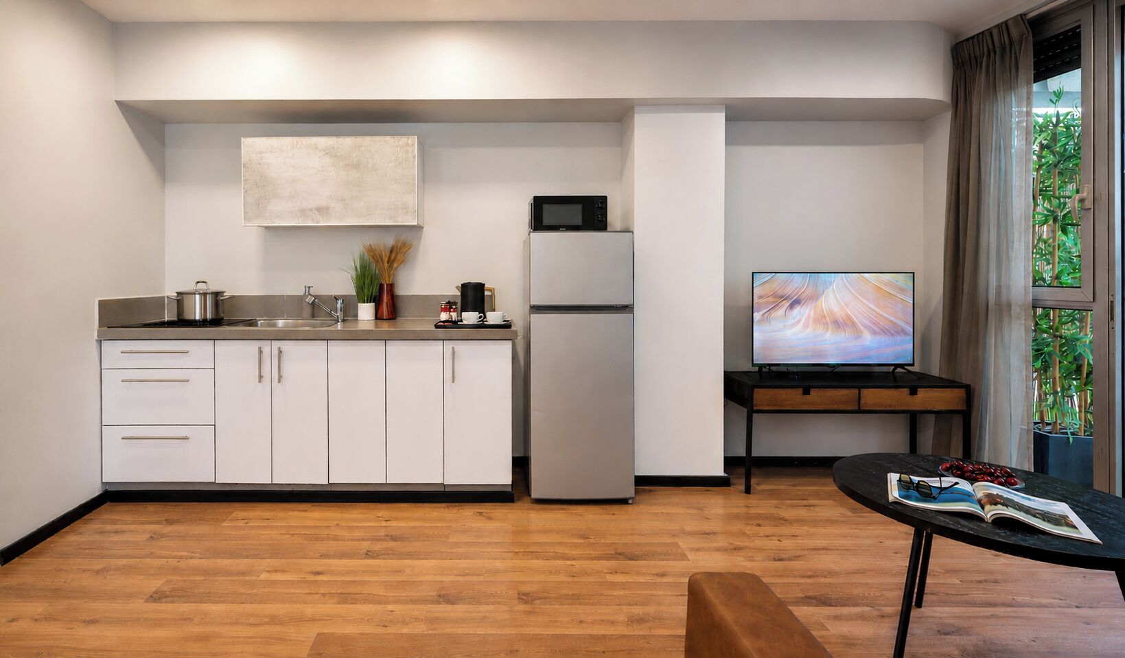 Eine moderne Küche mit weißen Schränken, einer Mikrowelle und einem Kühlschrank. Es gibt einen Essbereich mit Tisch und Stühlen sowie ein großes Fenster mit Vorhängen.