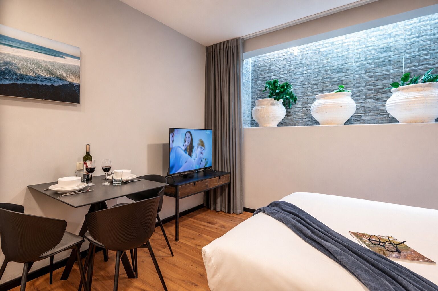 A hotel room with a bed, a desk with a television, a dining table, chairs, and a window with plants.
