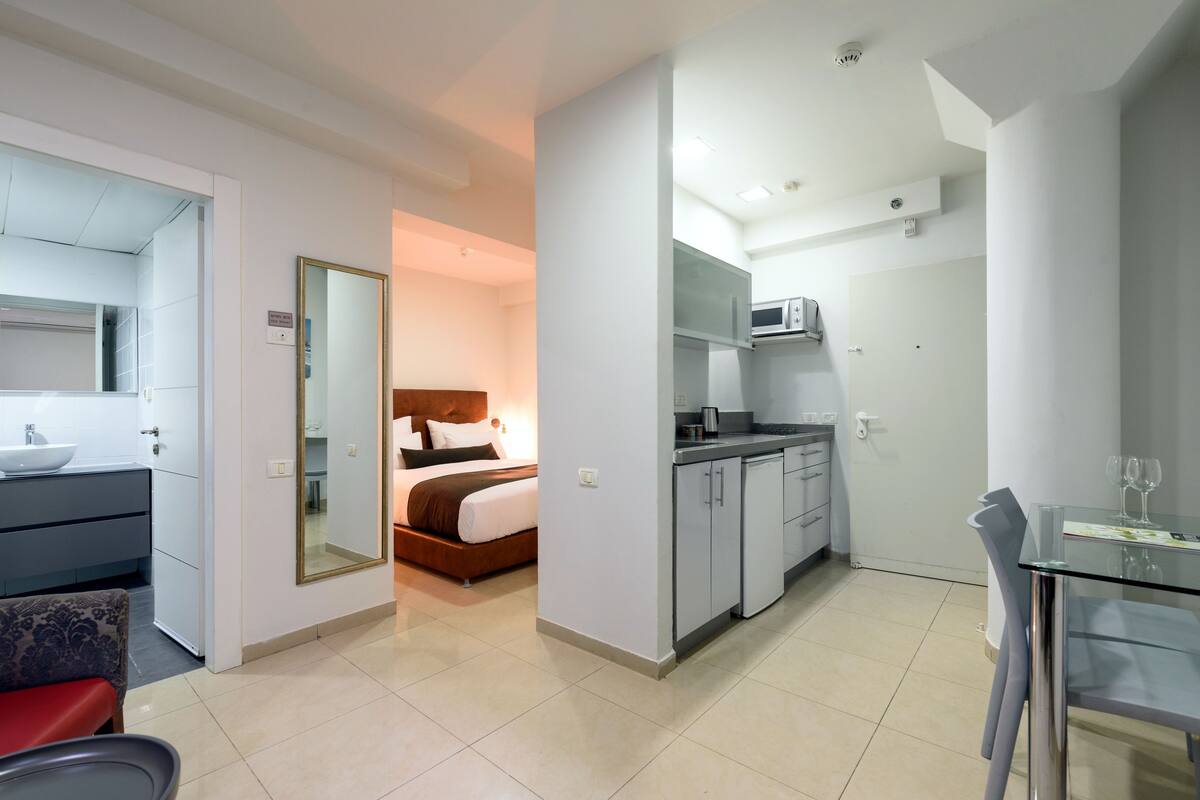 A modern hotel room with a bed, a bathroom, a kitchenette, and a dining area.