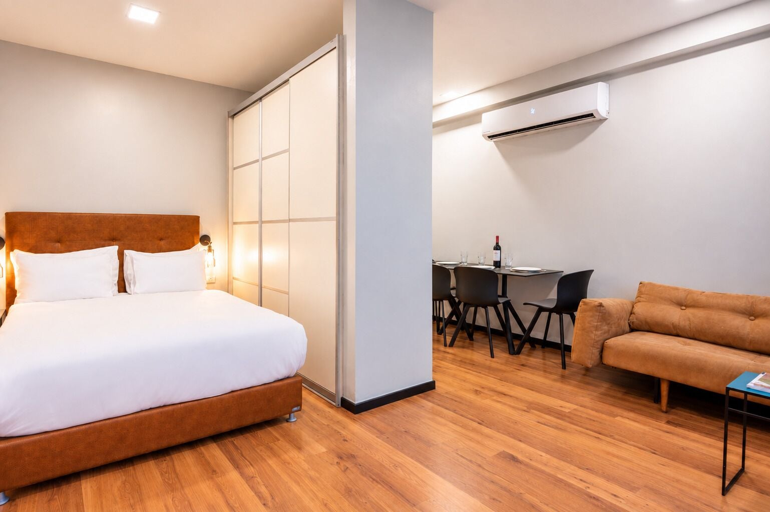 A hotel room with a bed, a desk with chairs, an air conditioner, and a sofa.