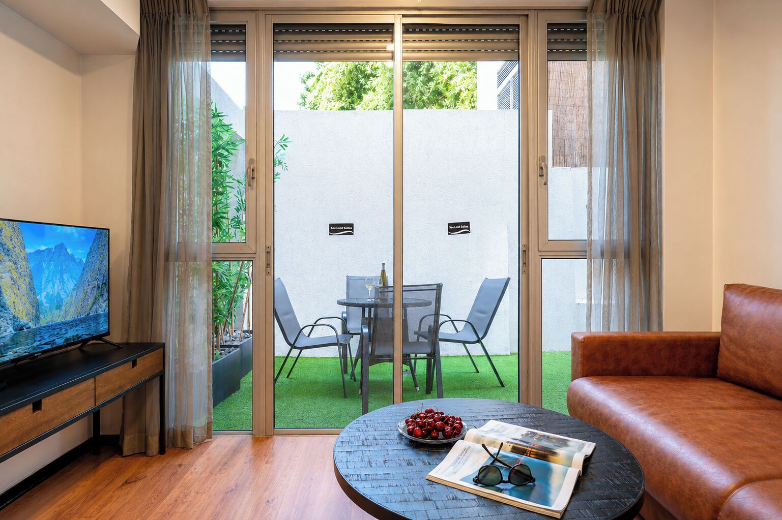 A modern living room with a television, a leather sofa, a coffee table with a magazine and sunglasses, and a dining area with chairs and a table.