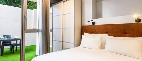 A hotel room with a bed, a nightstand, a table with chairs, and a sliding glass door leading to a green outdoor area.