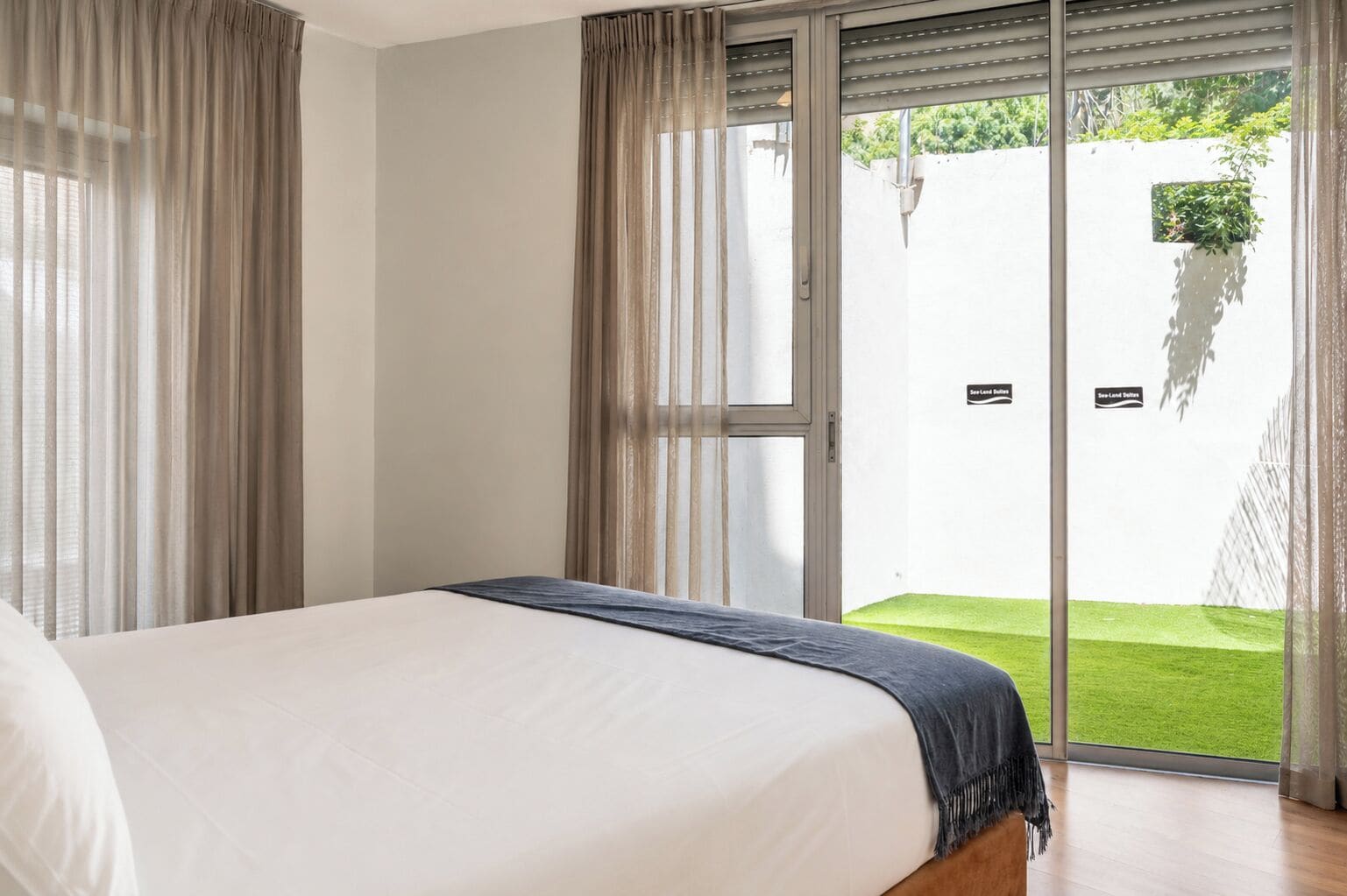 A bedroom with a bed, a view of a green lawn through a sliding glass door, and a white wall with a small window.