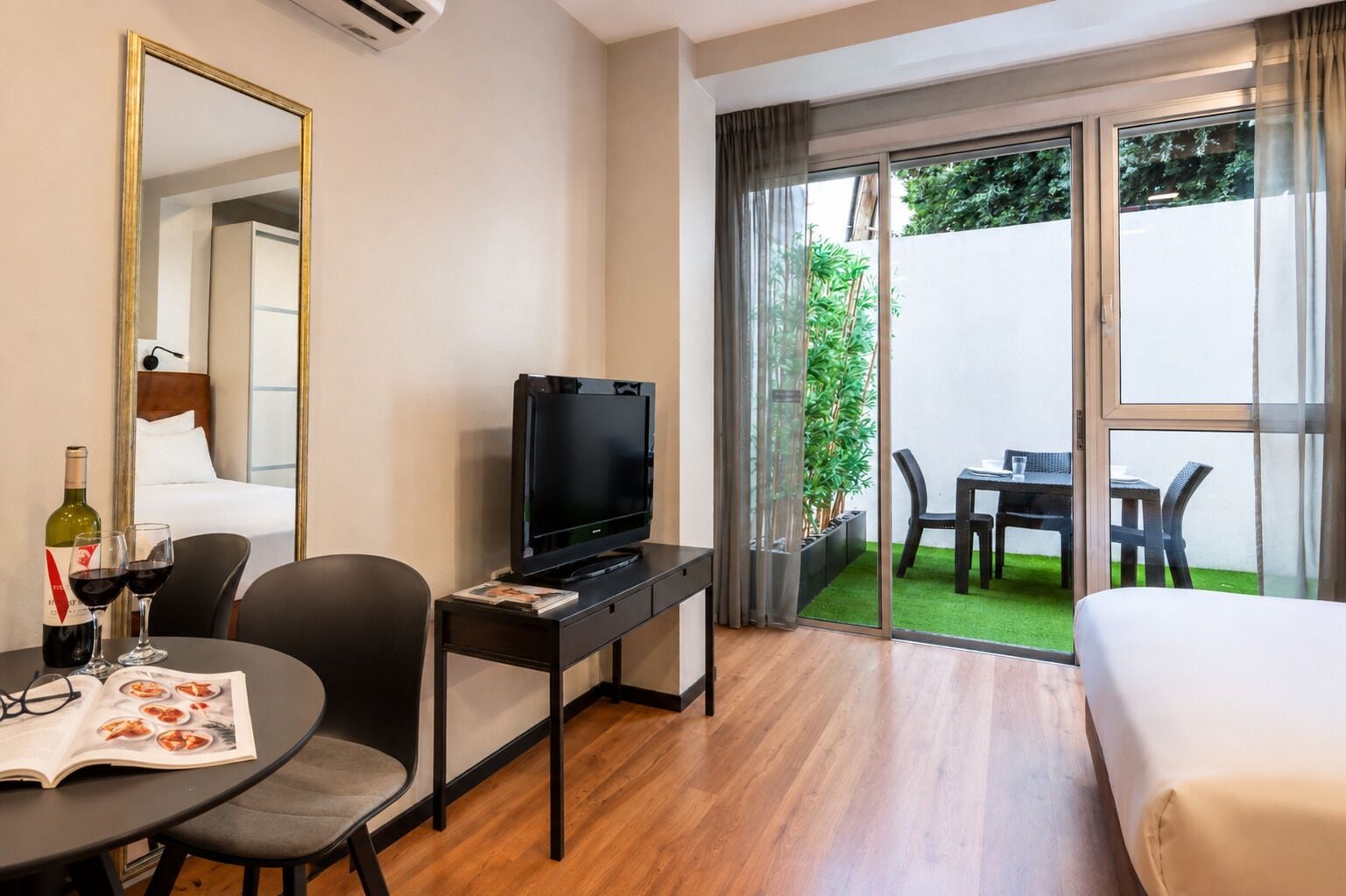 A hotel room with a bed, a desk with a television, a dining table with chairs, and a balcony with a table and chairs.