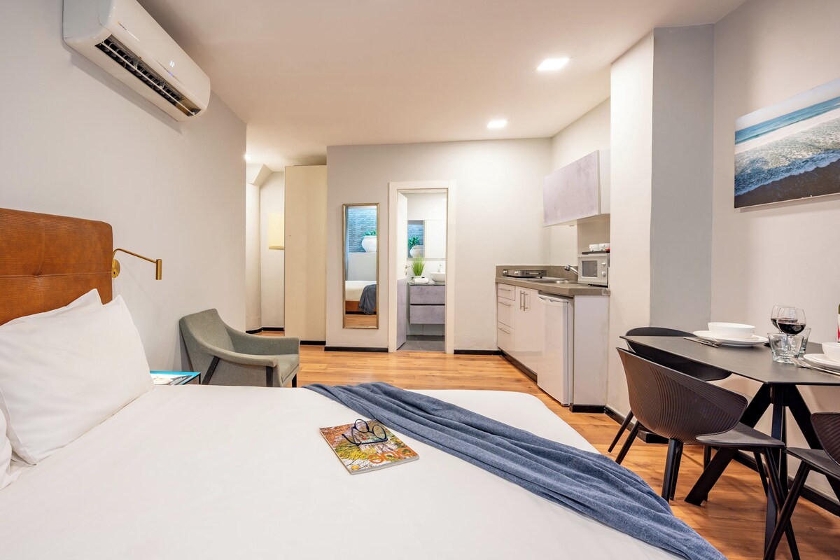 A hotel room with a bed, a desk, a chair, and a small kitchenette.