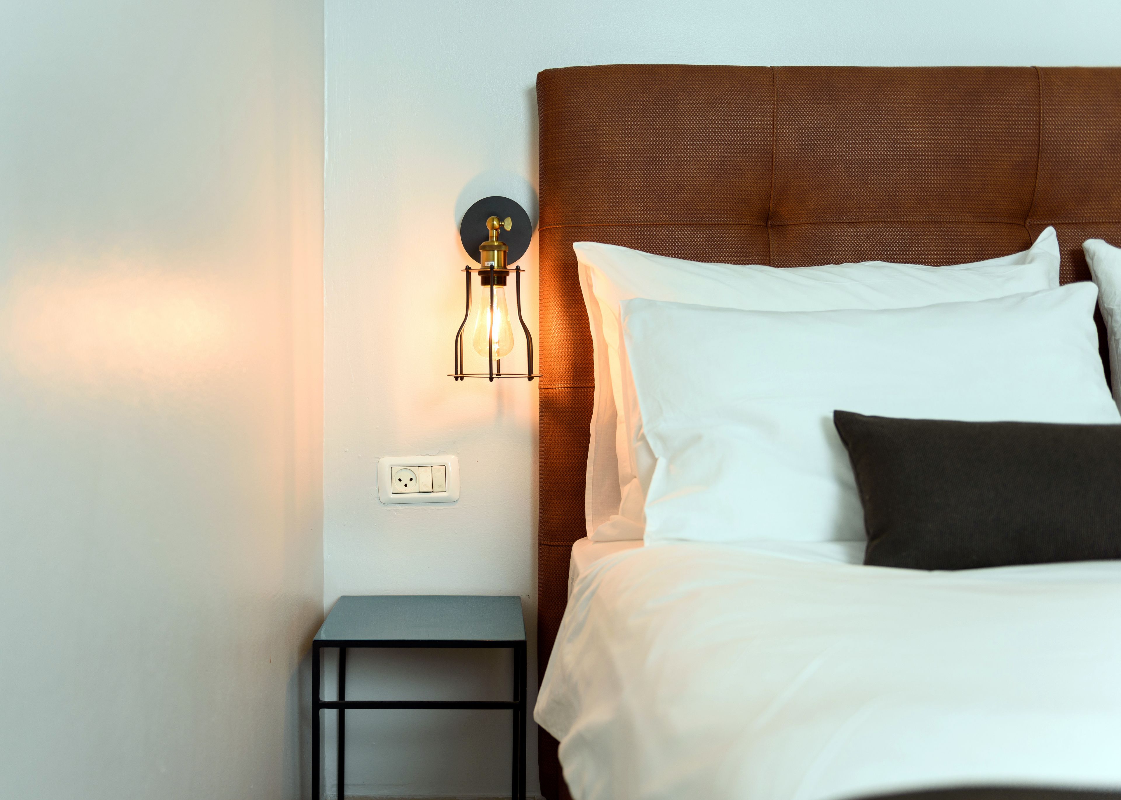 Un lit bien fait, avec des oreillers blancs et foncés, une petite table de chevet et un luminaire fixé au mur.