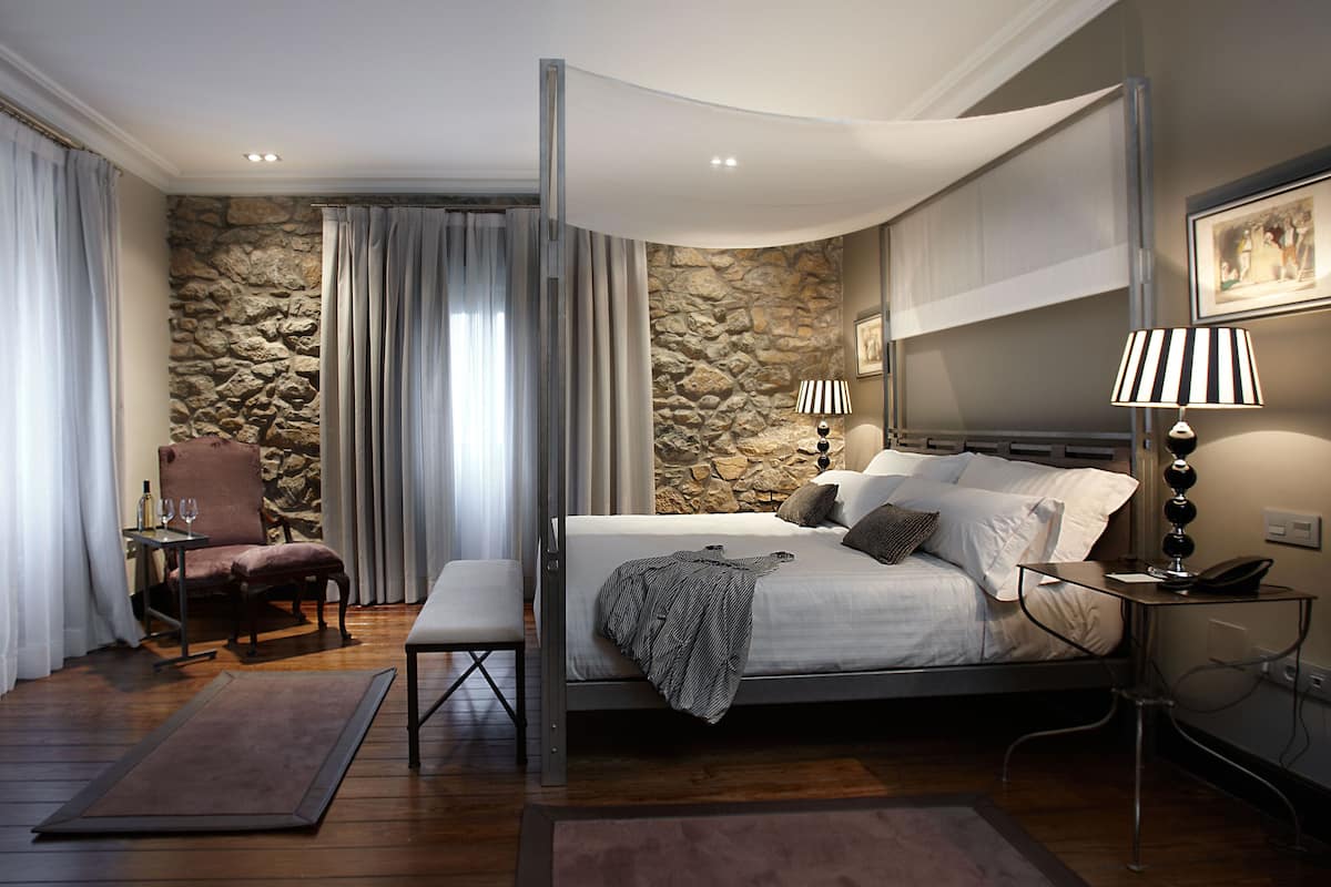 A modern bedroom with a stone wall, a bed with white linens, a chair, a table with wine glasses, and a lamp.