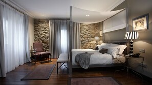 A modern bedroom with a stone wall, a bed with white linens, a chair, a table with wine glasses, and a lamp. - Hotel Iriarte Jauregia (Bidania-Goiatz)