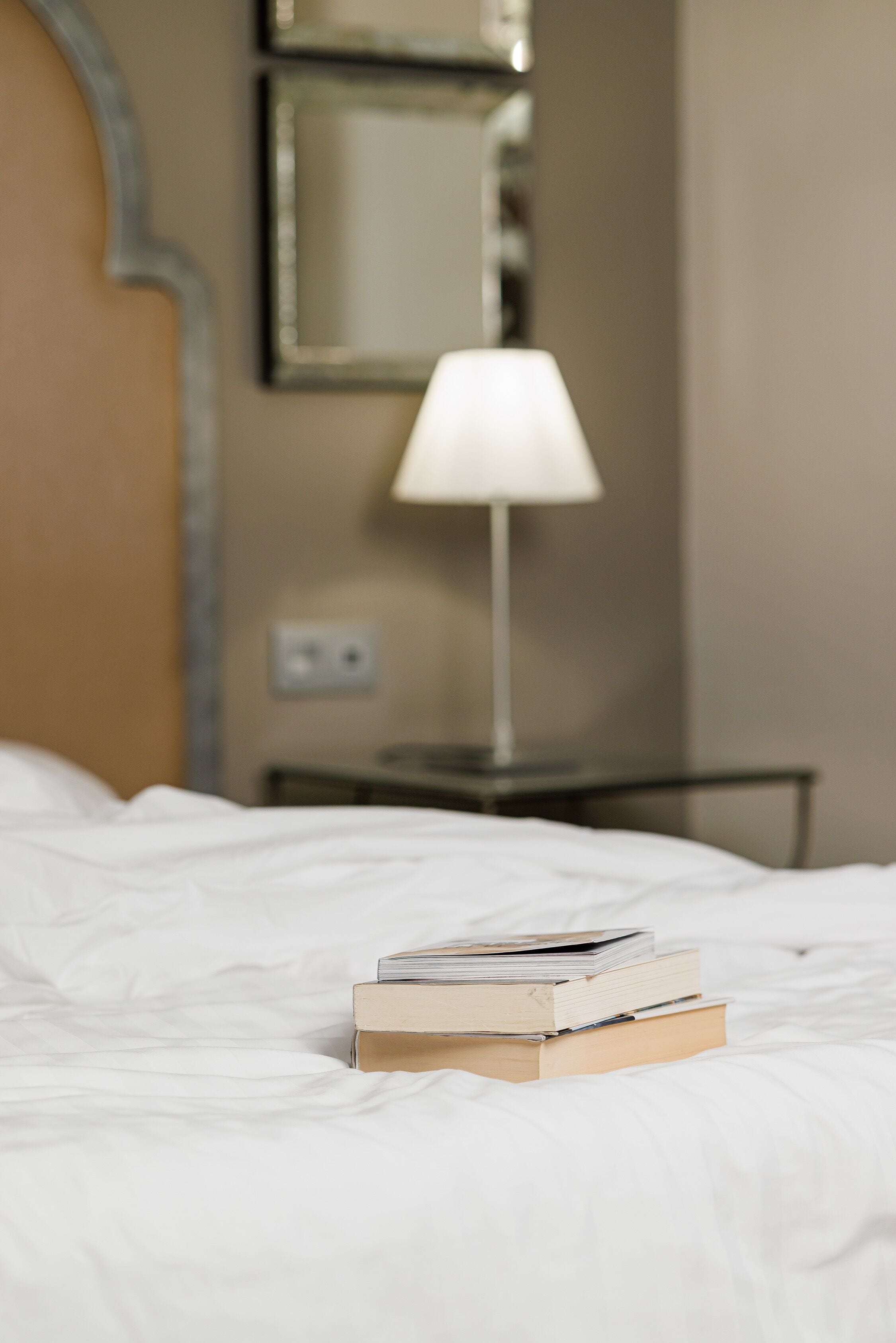 A neatly made bed with a stack of books on it, a bedside lamp, and a mirror on the wall.