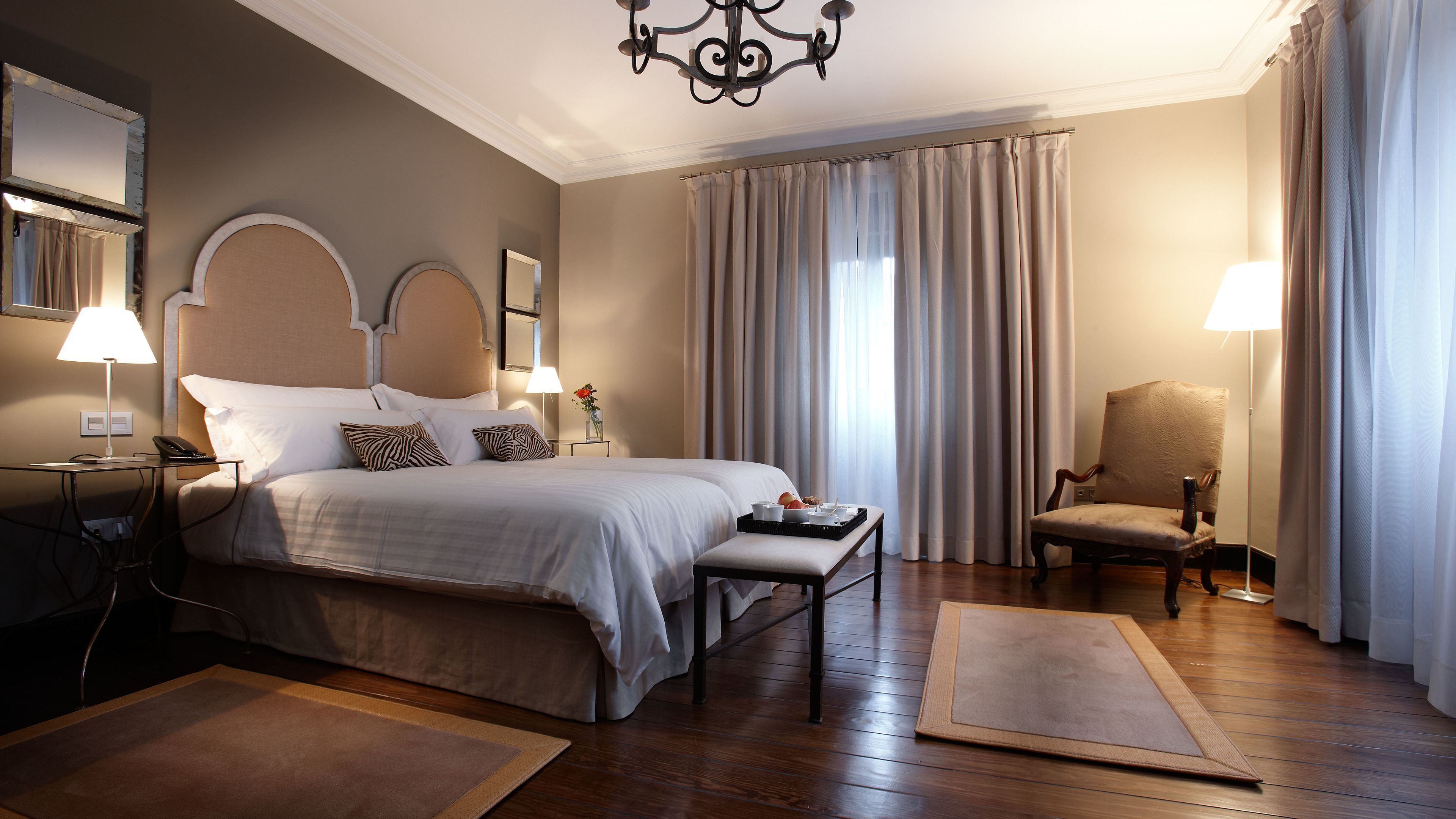 A hotel room with a large bed, bedside lamps, a chair, a desk, and a chandelier.
