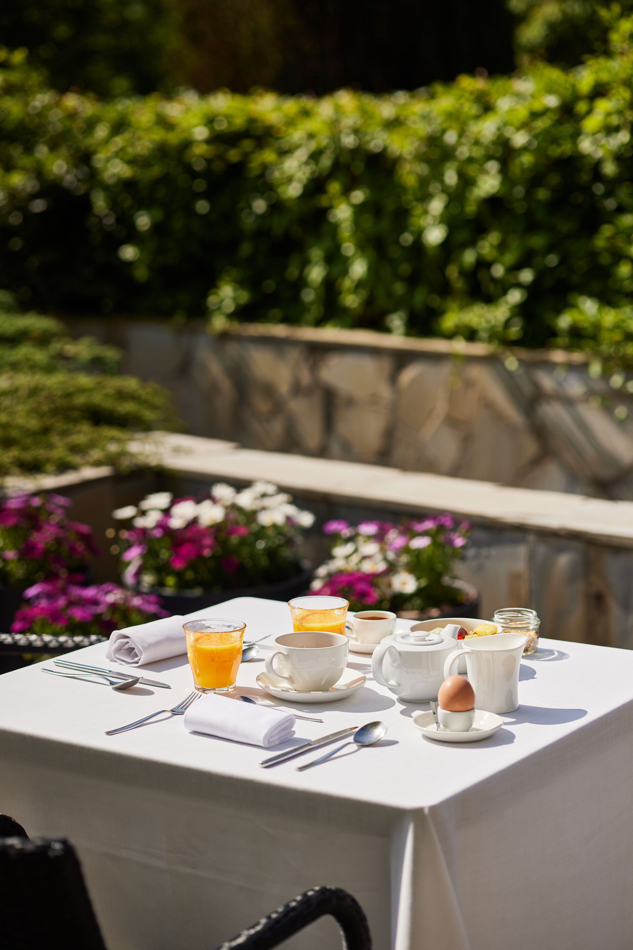 Une table dressée à l’extérieur, recouverte d’une nappe blanche, avec un verre de jus d’orange, une tasse, une théière et un pichet.