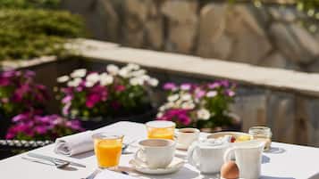 Une table dressée à l’extérieur, recouverte d’une nappe blanche, avec un verre de jus d’orange, une tasse, une théière et un pichet.