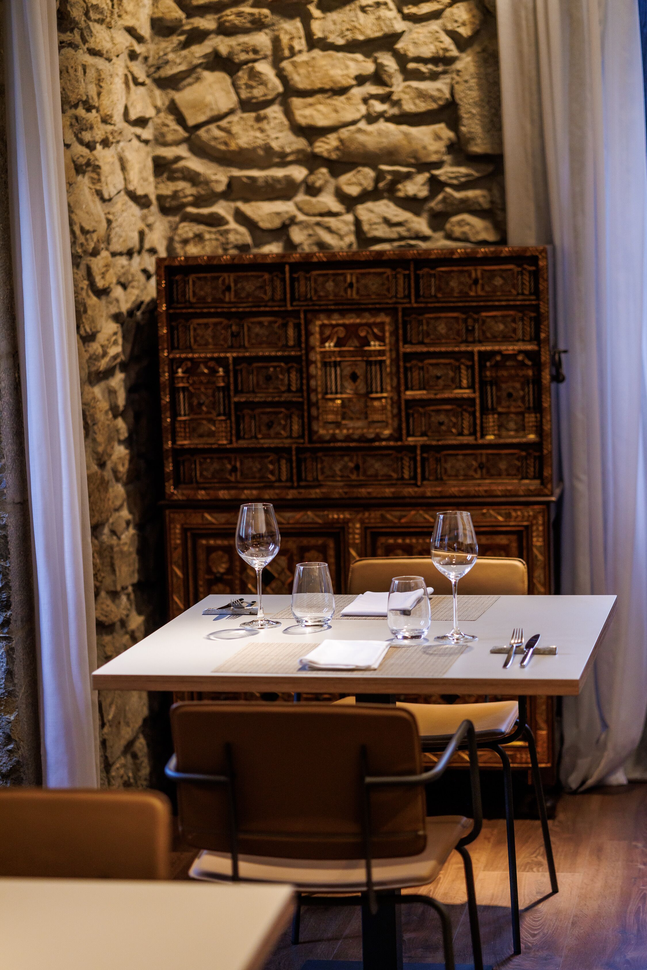 A dining table set with wine glasses, a menu, and cutlery, against a stone wall and wooden panel backdrop.