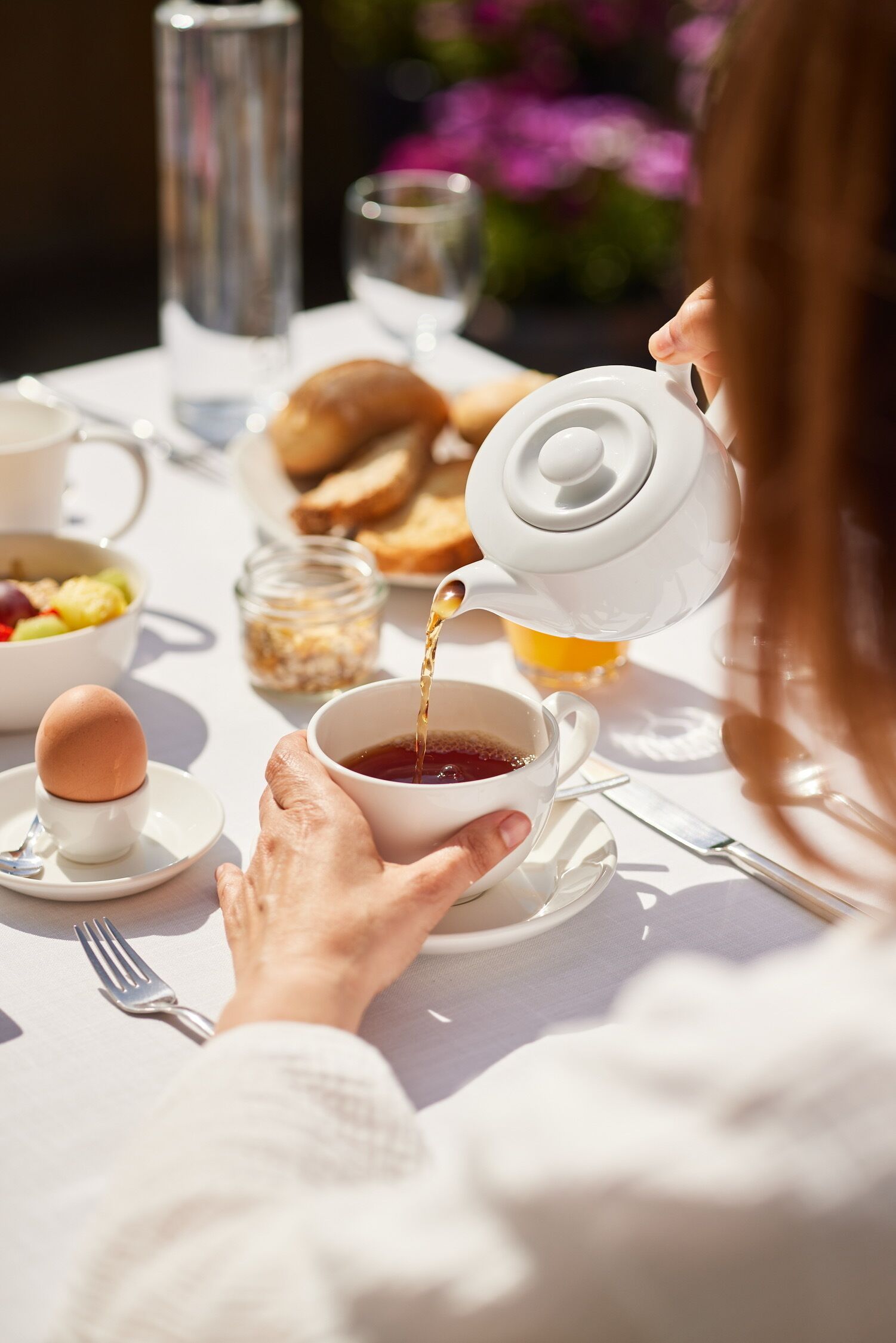 Un desayuno con una tetera sirviendo té en una taza, un bol de fruta y un plato de cruasanes.