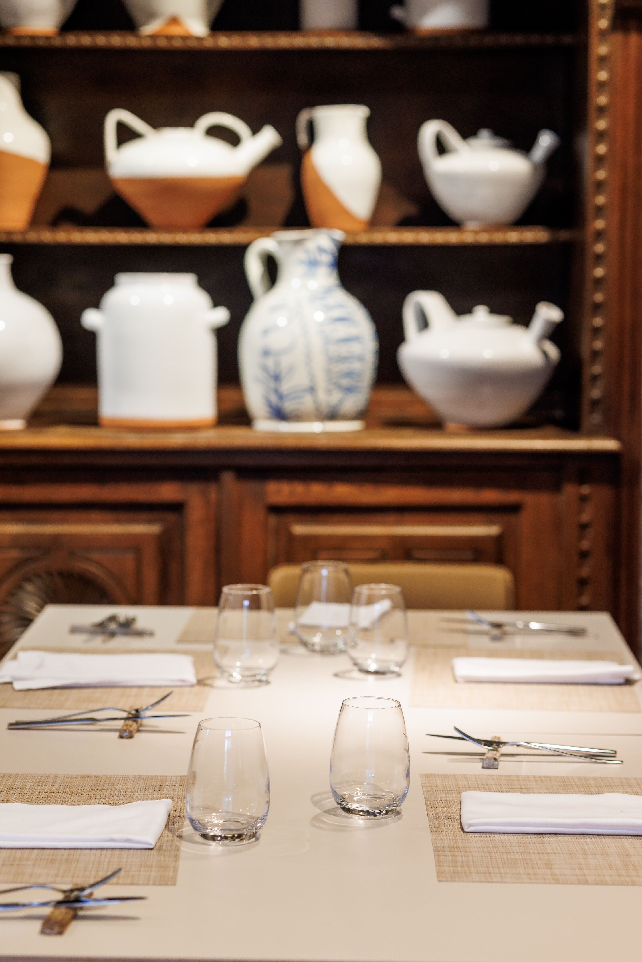 A dining table set with plates, cutlery, and glasses, with a shelf of white and blue ceramic kitchenware in the background.
