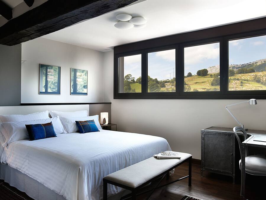 A bedroom with a large bed, a desk with a computer, and a window with a view of a green landscape.