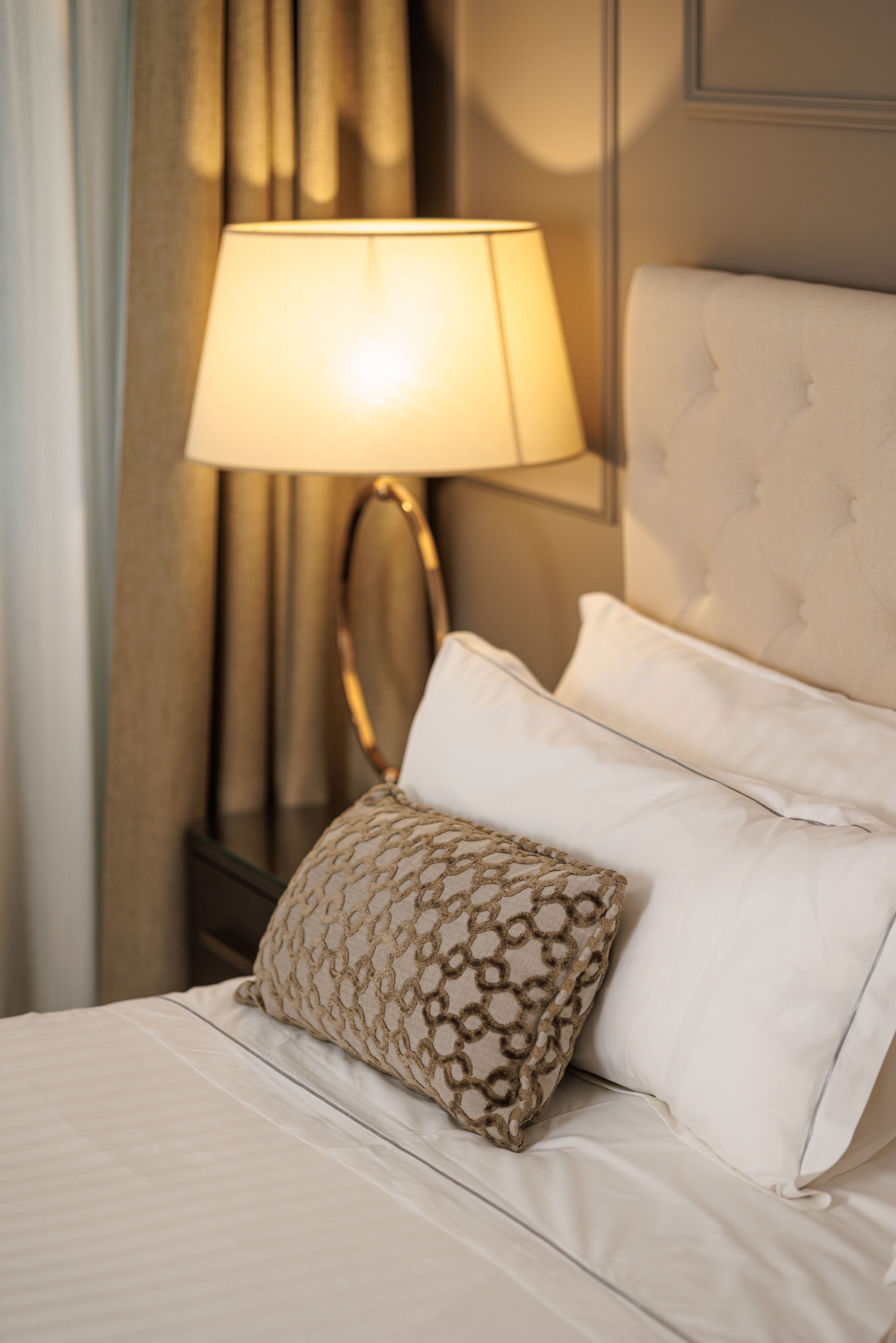 Una cama bien tendida con sábanas blancas y un cojín decorativo estampado, una lámpara de noche y un cabecero.