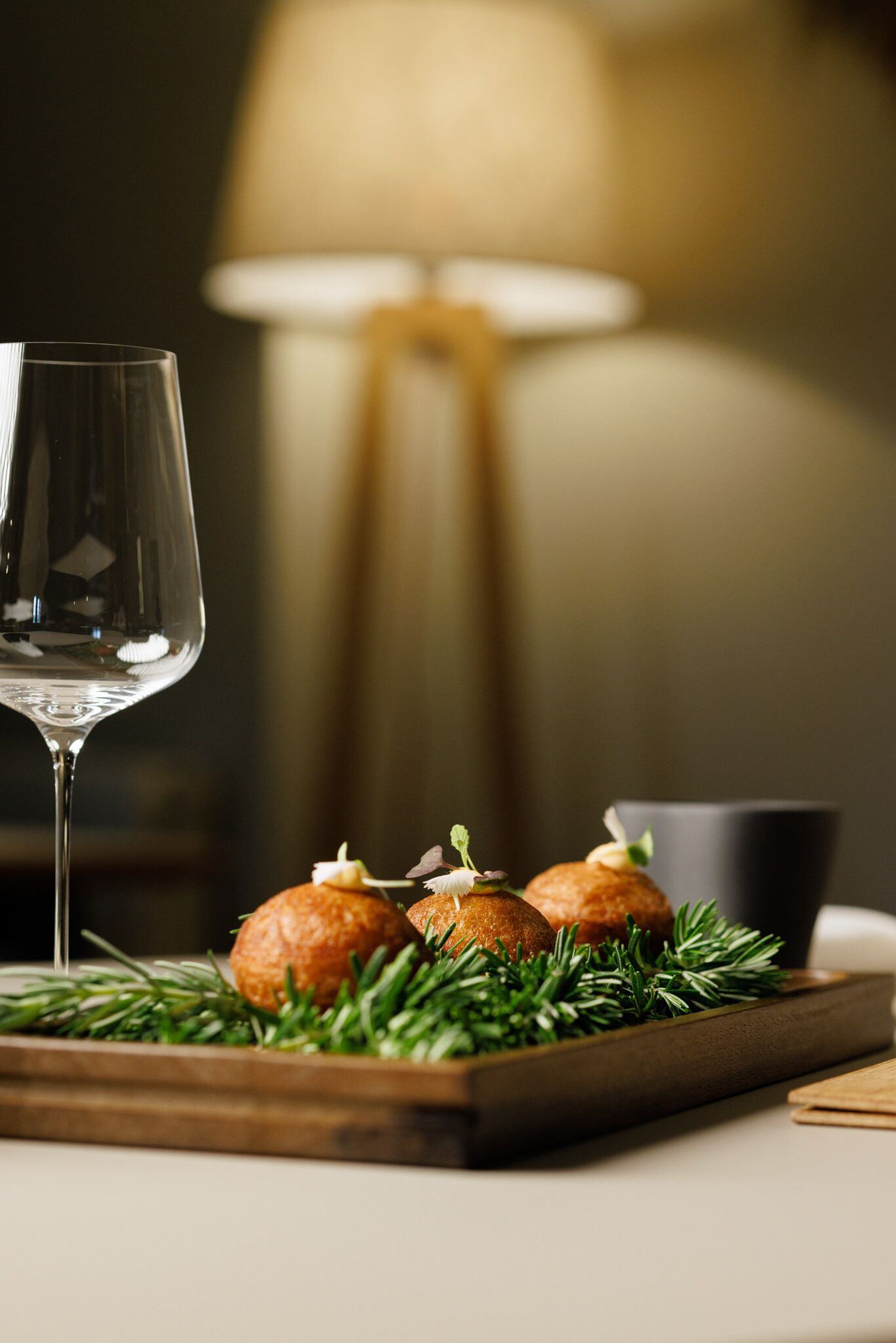 Een glas wijn, twee ronde gebakjes op een houten plank met groen, en een lamp op de achtergrond.