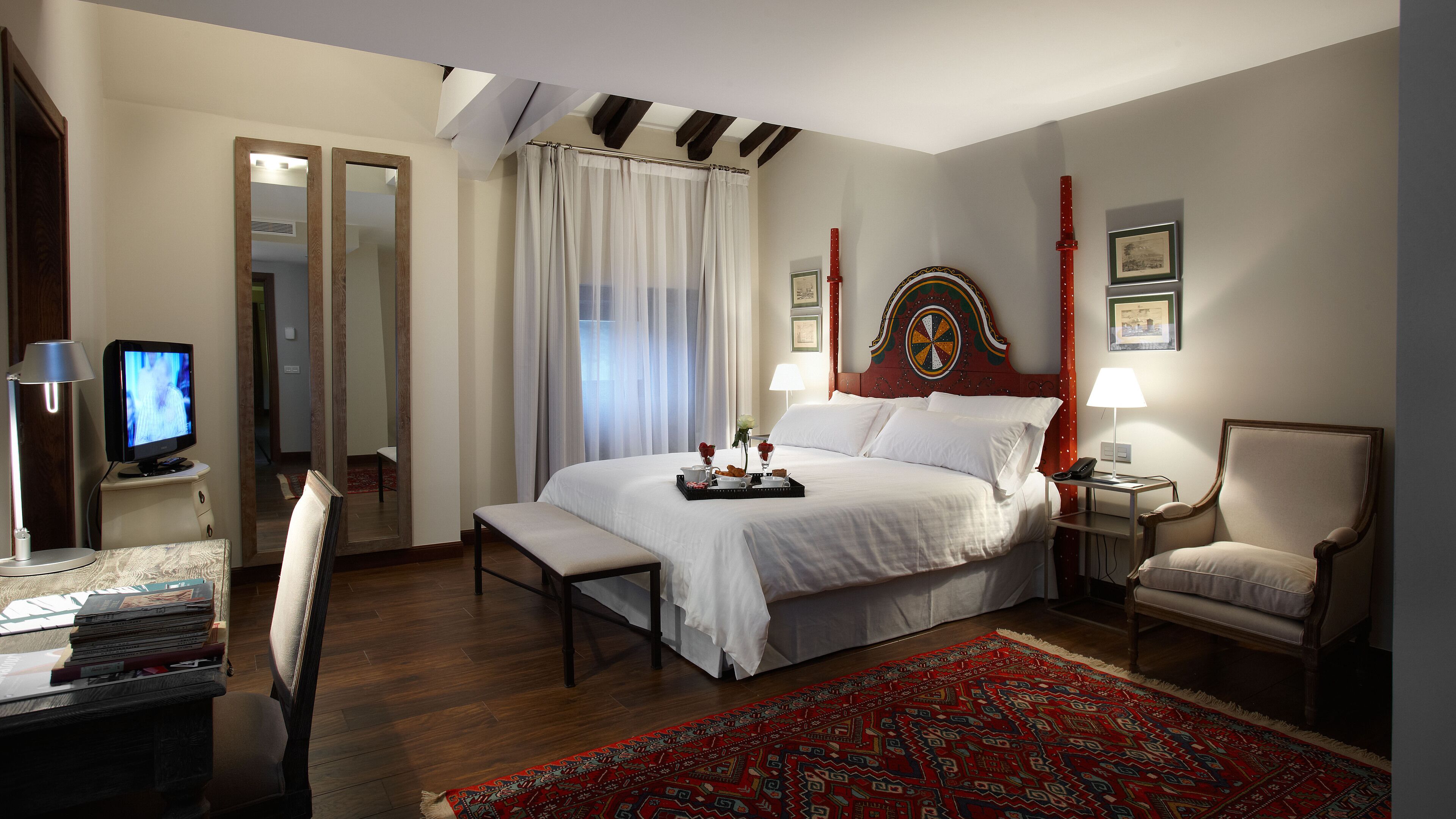 Habitación de hotel con una cama grande, un escritorio con computadora, una silla y una alfombra roja estampada.