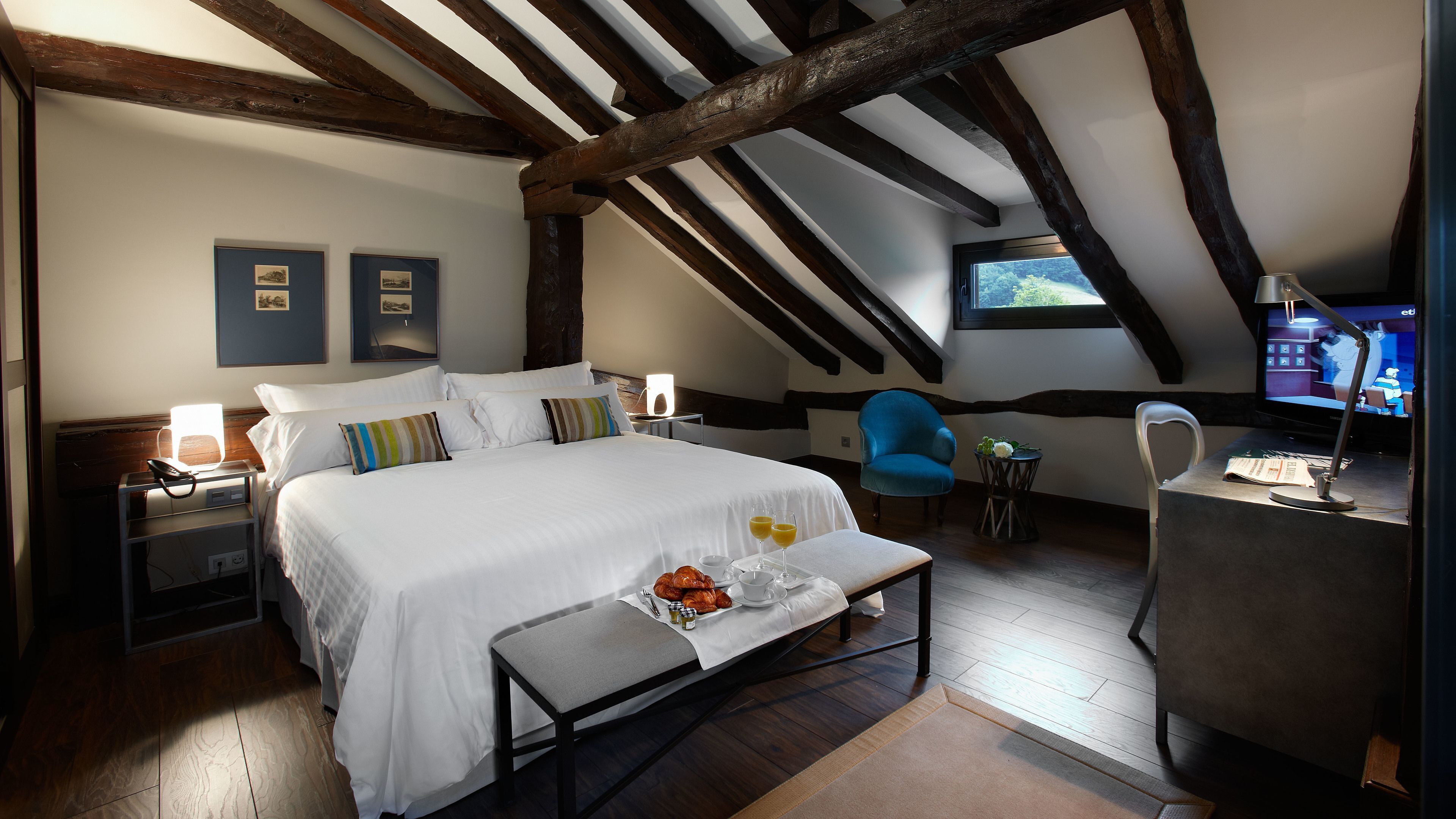 A bedroom with a large bed, bedside tables, a chair, a small table with a tray of breakfast, and a television.