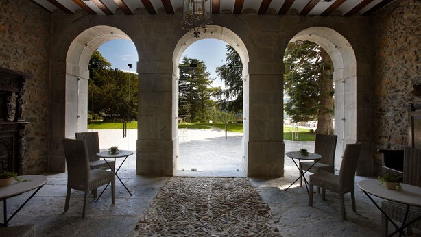Una habitación con paredes de piedra y tres aberturas en arco, mobiliario de mimbre y vistas a un parque.