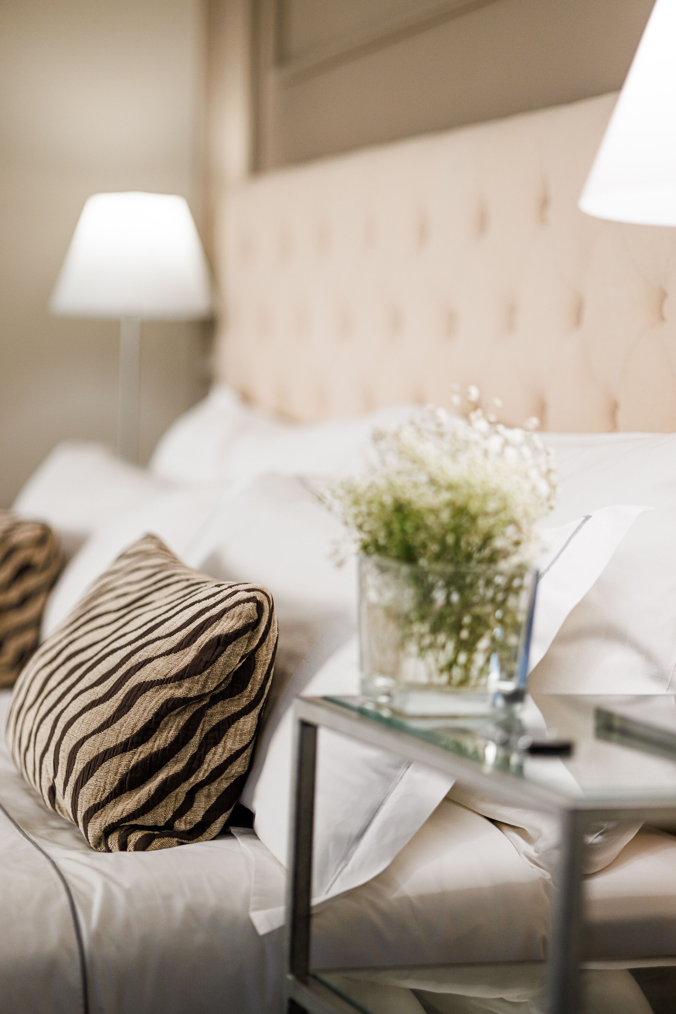 A neatly made bed with striped pillows, a glass bedside table, and a vase with flowers.
