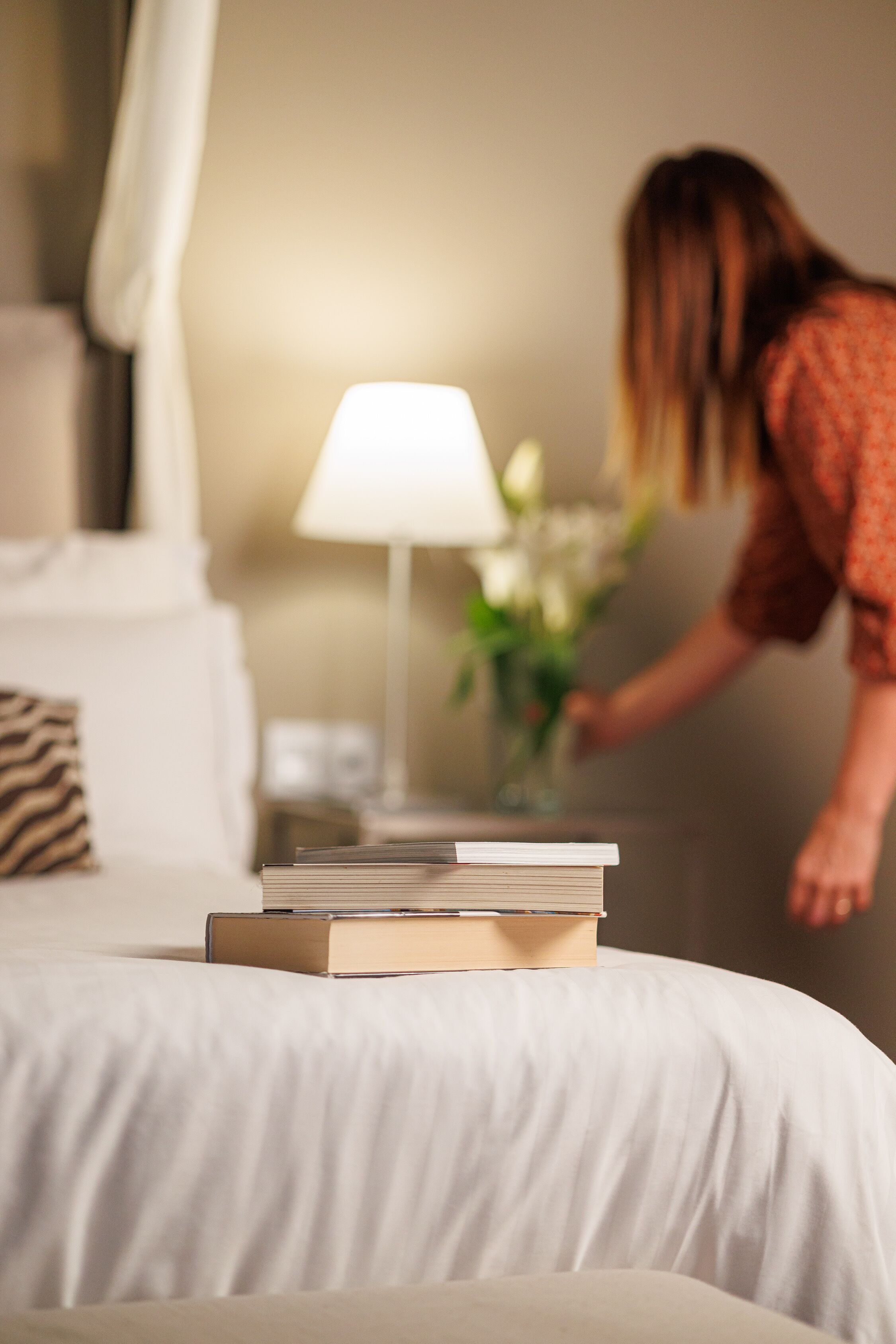 A neatly made bed with a stack of books, a bedside lamp, and a vase with flowers.
