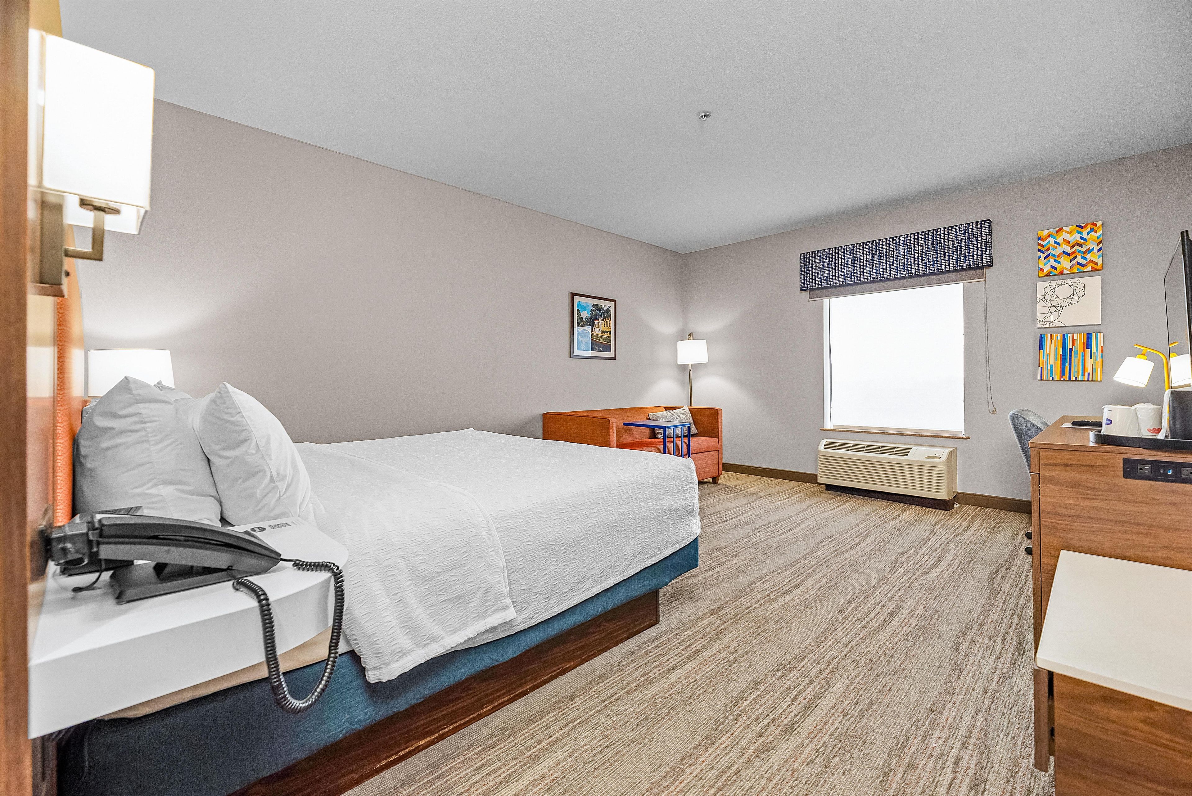 A hotel room with a bed, bedside table, lamp, desk, chair, and a window with a view.