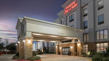 Um Hampton Inn & Suites com entrada coberta, pilares de pedra e céu claro.