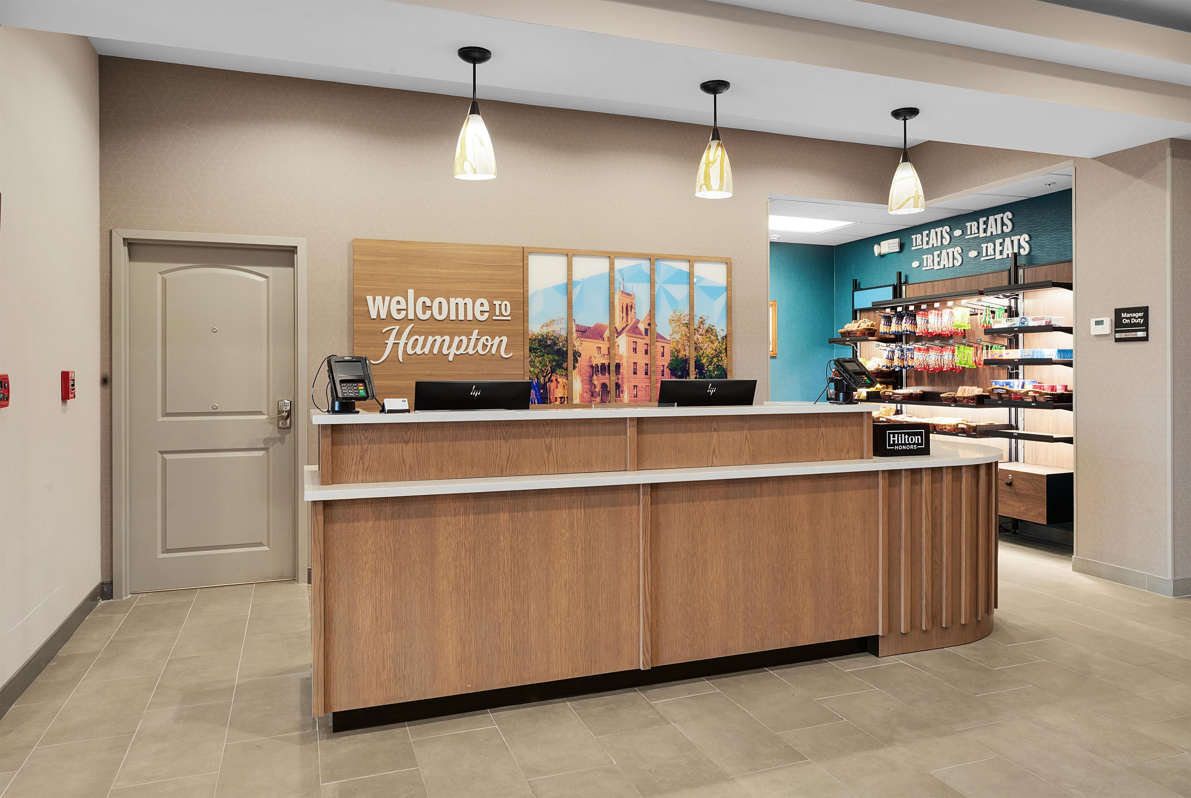 A hotel reception desk with a 'Welcome to Hampton' sign, a digital display, and a 'Treats Treats Treats' snack area.