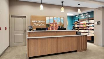 A hotel reception desk with a 'Welcome to Hampton' sign, a digital display, and a 'Treats Treats Treats' snack area.