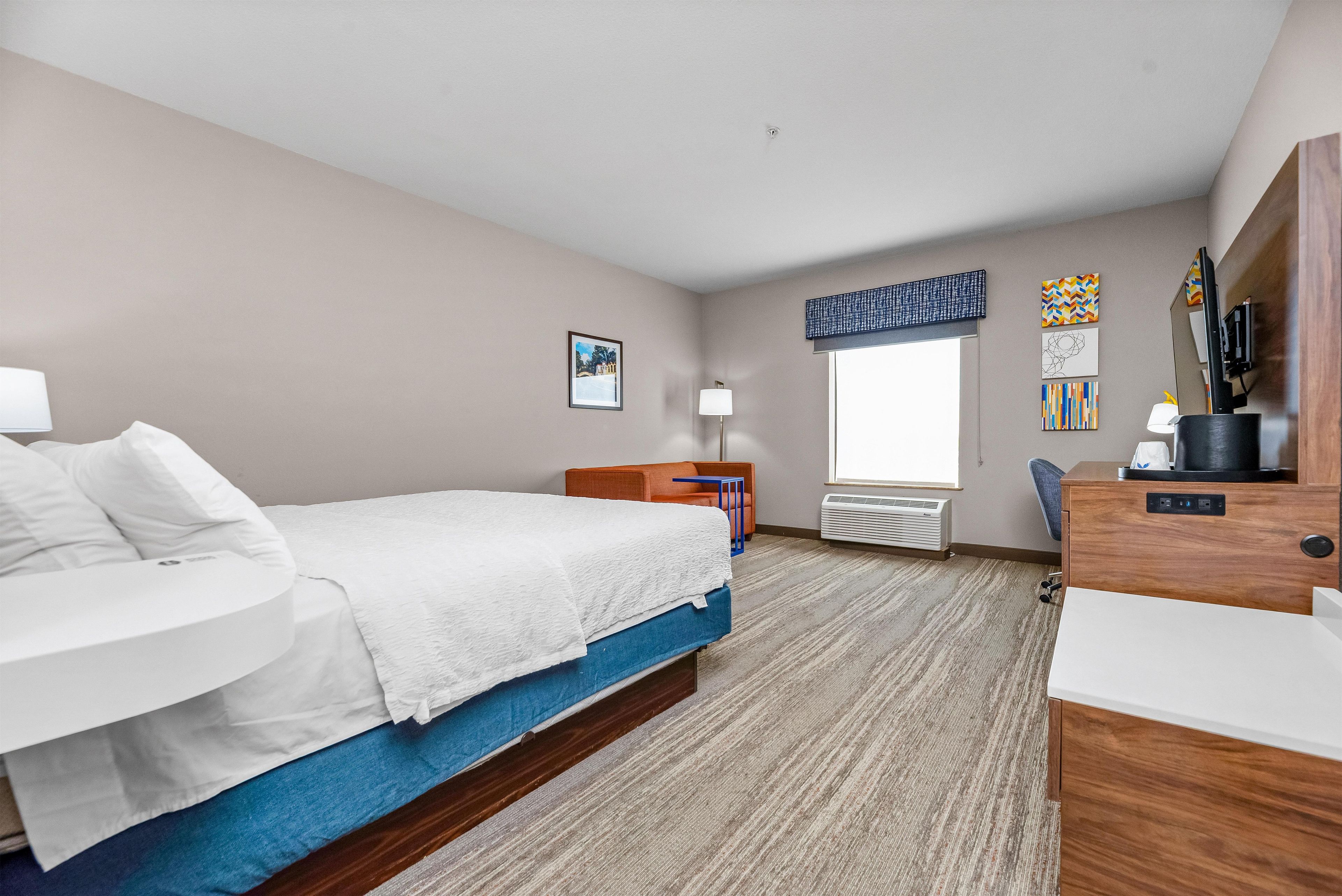 A hotel room with a bed, a desk, a chair, a window, and artwork on the wall.