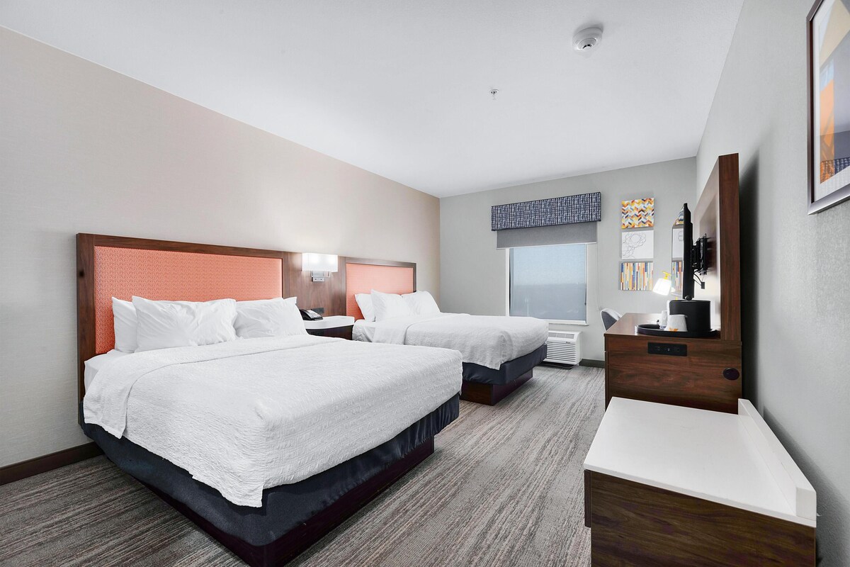 A hotel room with two beds, a desk, a chair, a large window, and a view of the city.