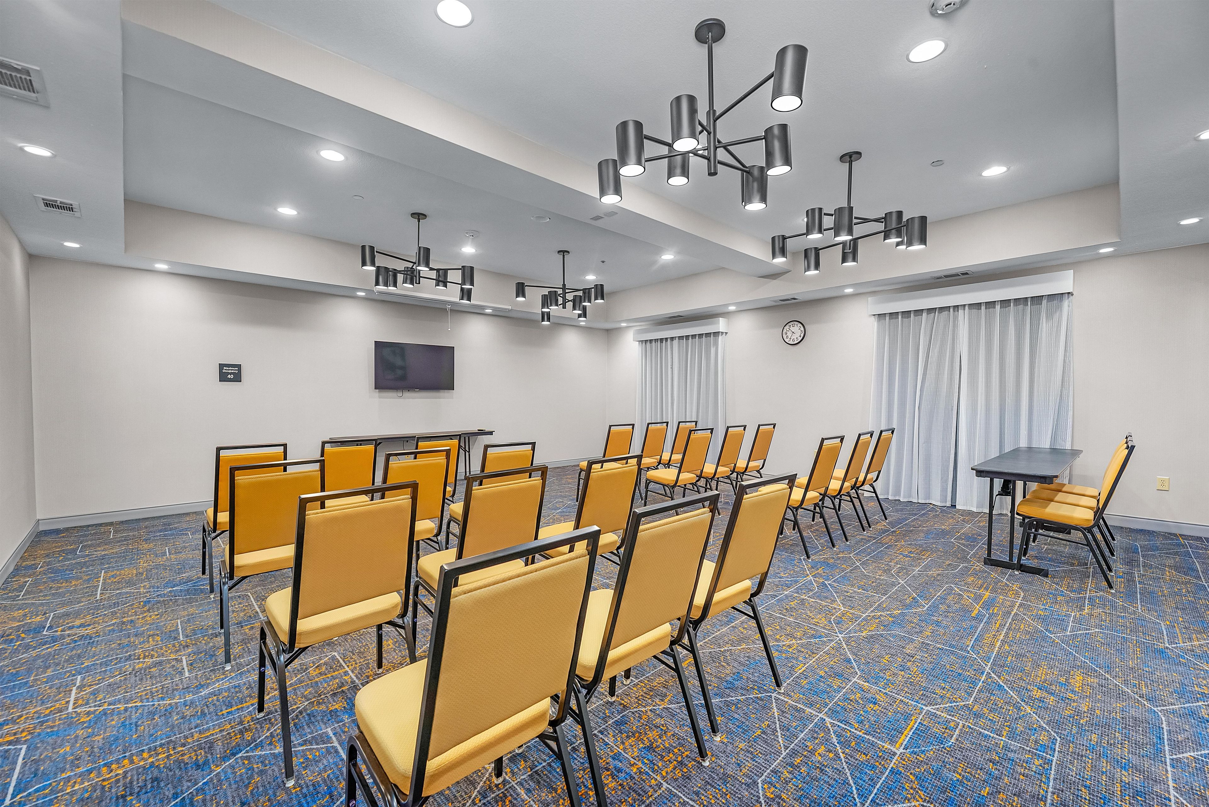 A conference room with rows of yellow chairs, a black table, a clock on the wall, and a ceiling light fixture.