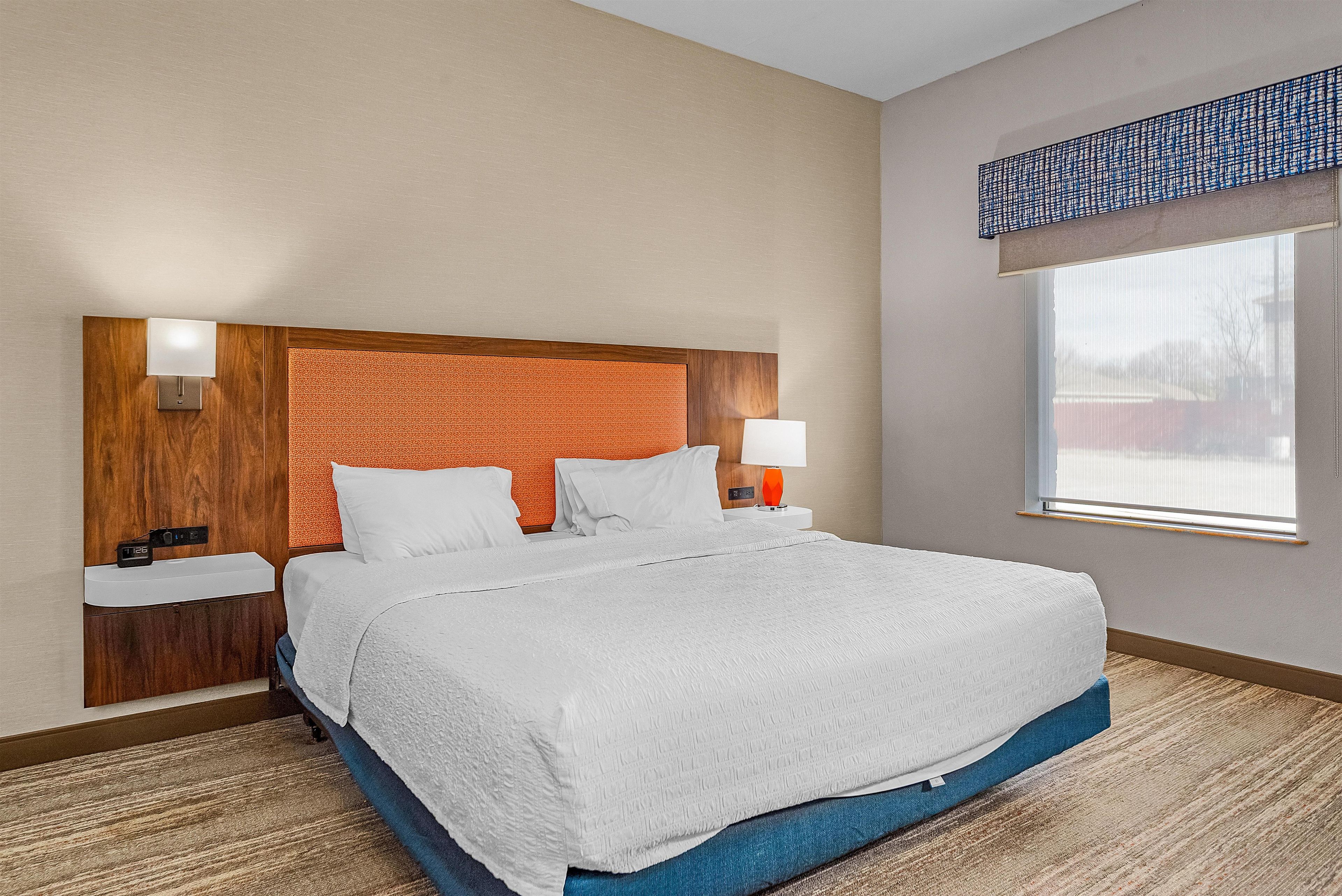 A hotel room with a large bed, two bedside tables, a wooden headboard, and a window with blinds.