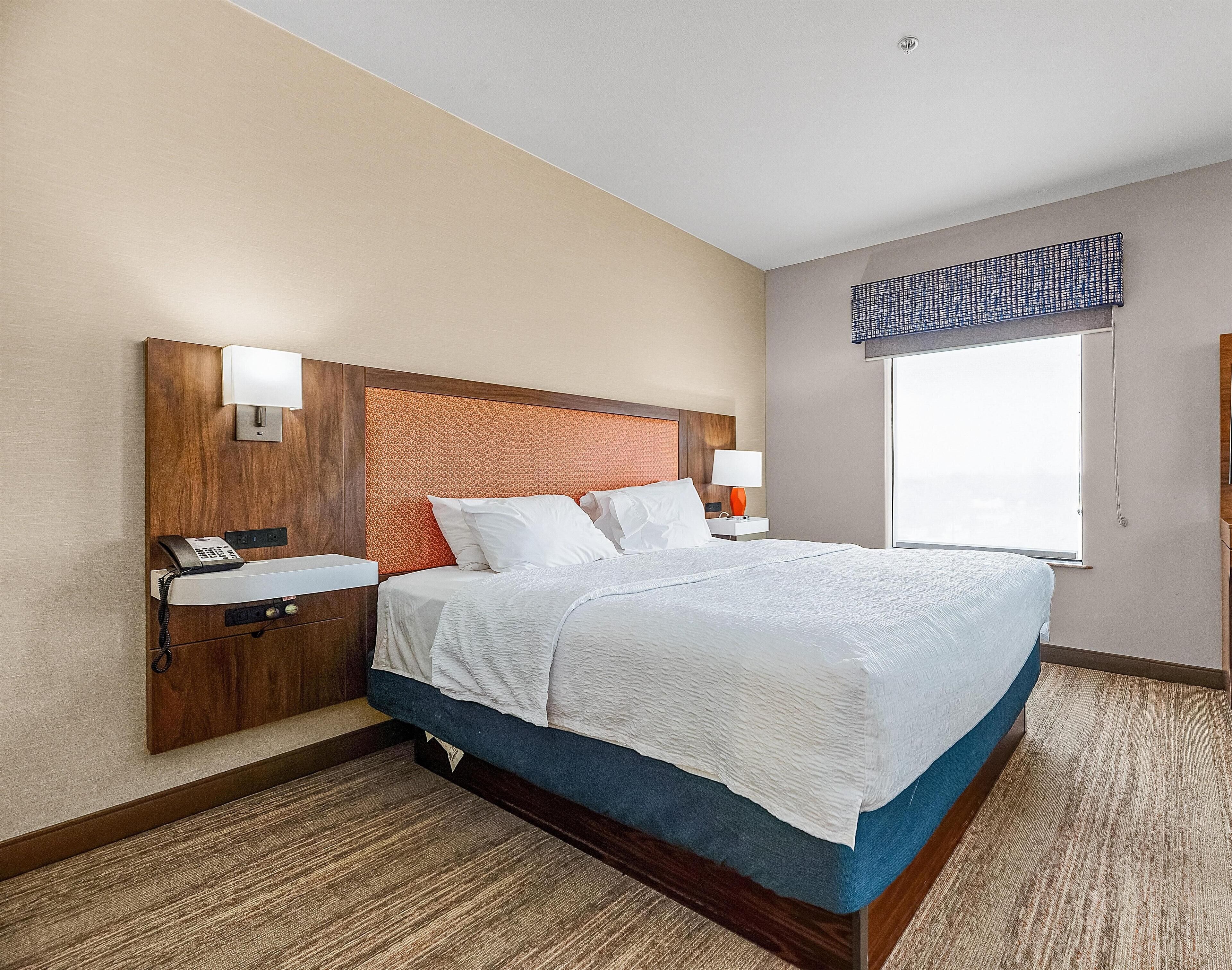 Habitación de hotel con cabecera de madera, cama con ropa blanca, mesita de noche con lámpara, teléfono y ventana con vistas.
