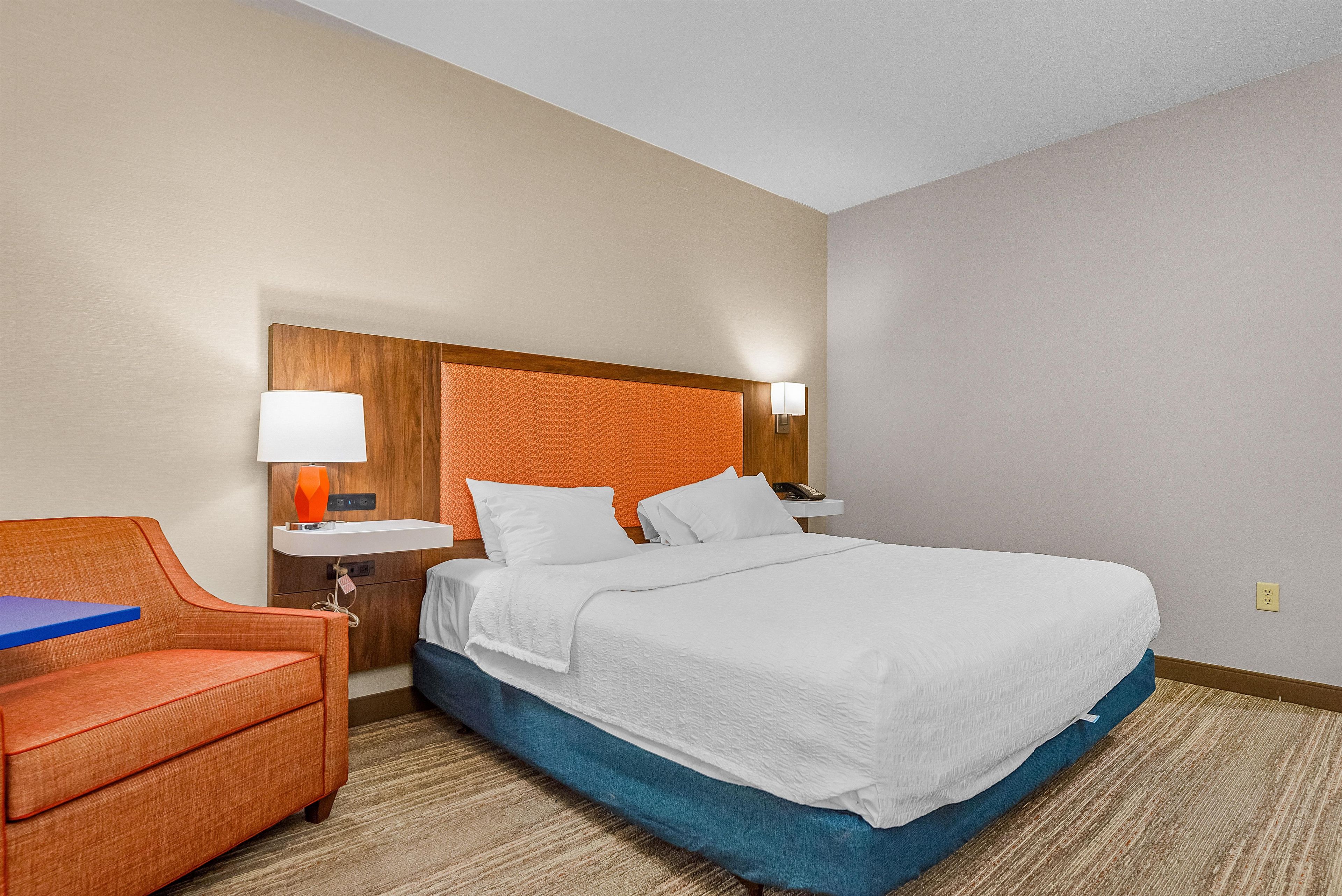 A hotel room with a bed, a nightstand, a lamp, a chair, and a wall-mounted phone.