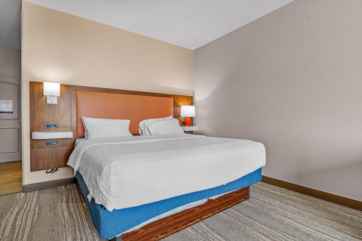 A hotel room with a large bed, a wooden headboard, a nightstand with a lamp, and a small table with a chair.