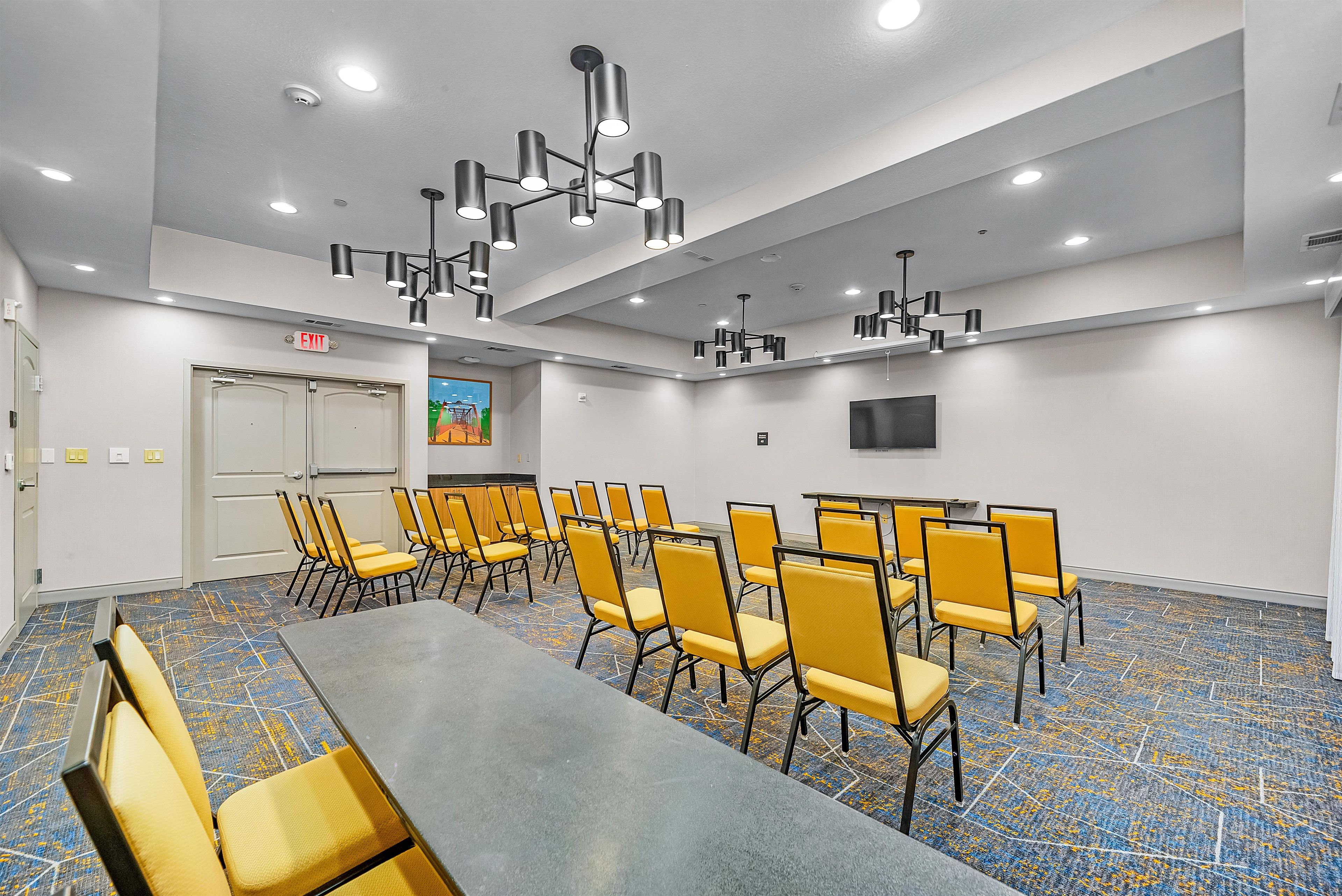 Sala de conferências com uma mesa comprida e várias cadeiras, luminárias no teto e sinal de saída.