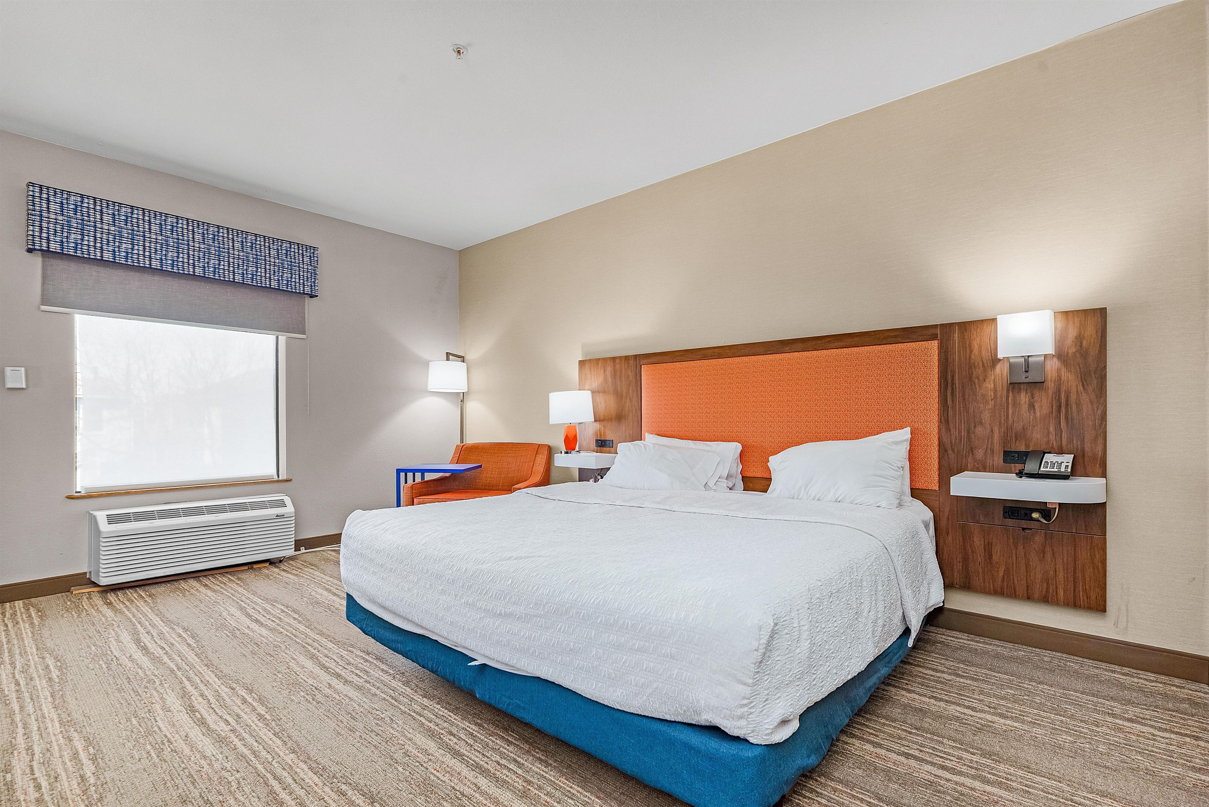 A hotel room with a large bed, a desk, a chair, a window, and a wall-mounted nightstand.